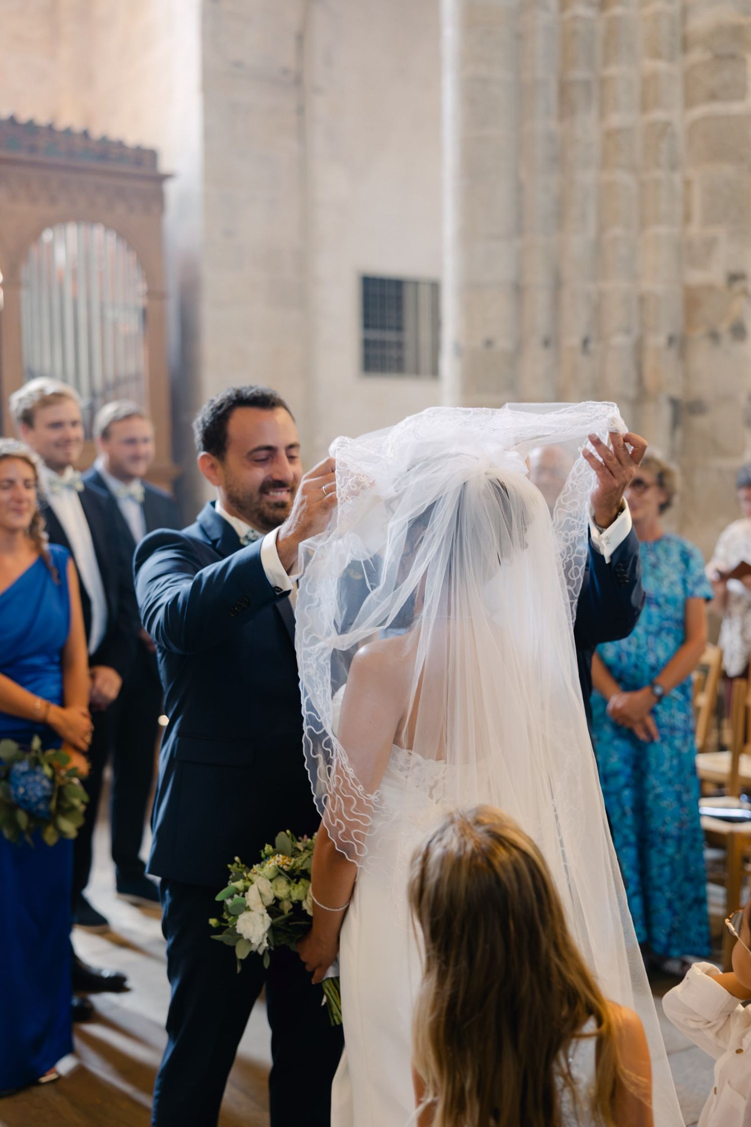 Groom lifting bride's lace veil during church wedding ceremony while guests watch in the background.