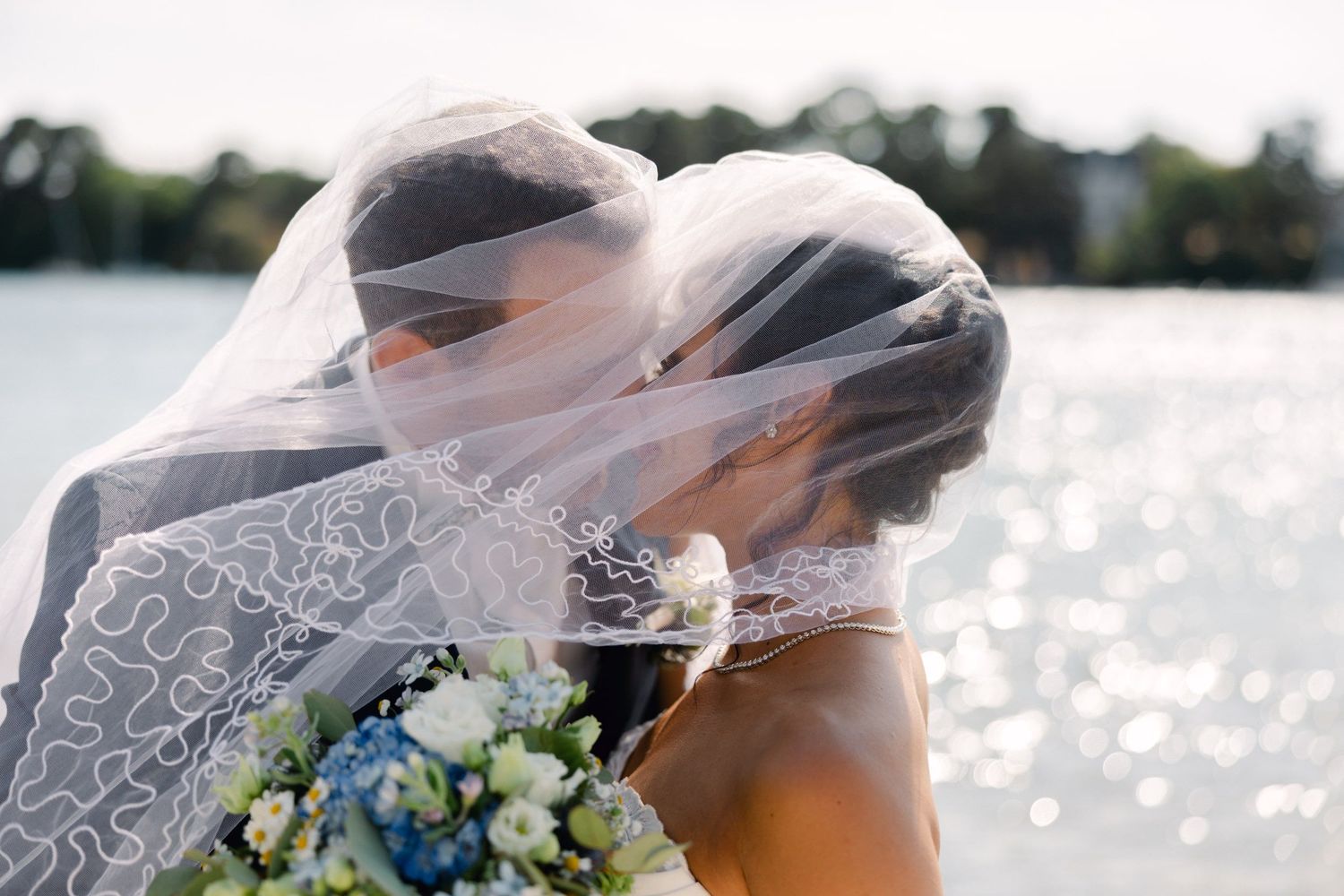 Bride and groom kissing under a white lace wedding veil by a shimmering lake, bride holding a blue floral bouquet.