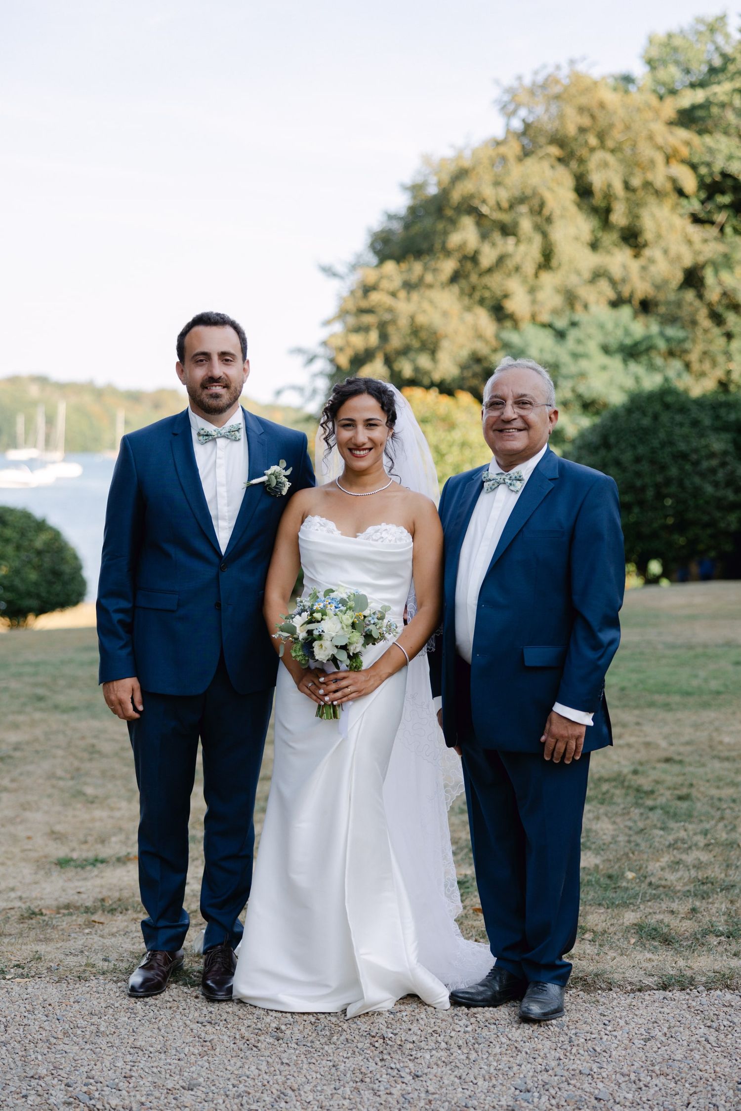 Bride in white gown with bouquet poses with two men in navy suits at an outdoor wedding ceremony.