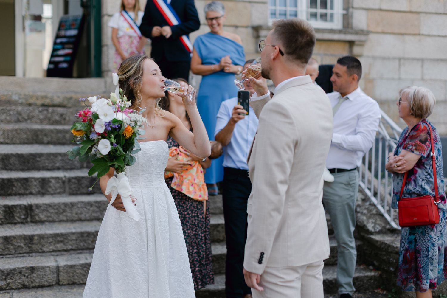 La mariée et le marié trinquent avec des verres à champagne sur des marches en pierre à l'extérieur de mairie après la cérémonie.
