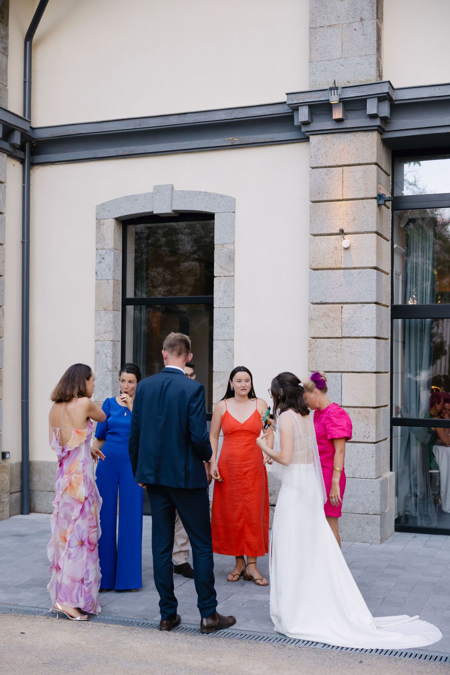 La mariée en robe blanche accueille les invités de mariage habillés de manière colorée à l'extérieur d'un élégant bâtiment en pierre.