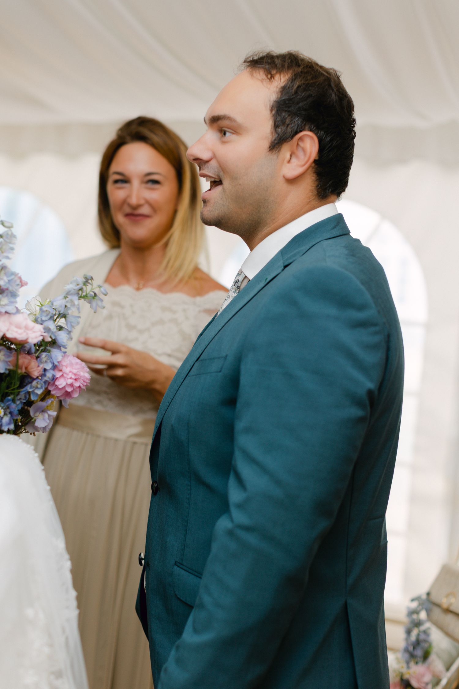 Marié souriant en costume sarcelle lors d'une cérémonie de mariage avec des arrangements floraux et des invités en arrière-plan.