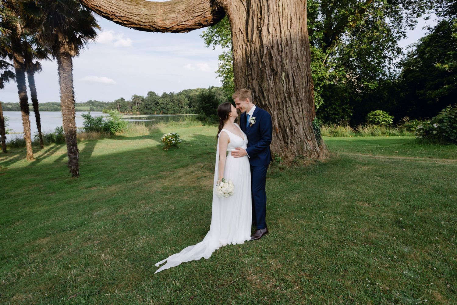 La mariée et le marié partagent un moment romantique près d'un grand arbre au bord du lac le jour du mariage.
