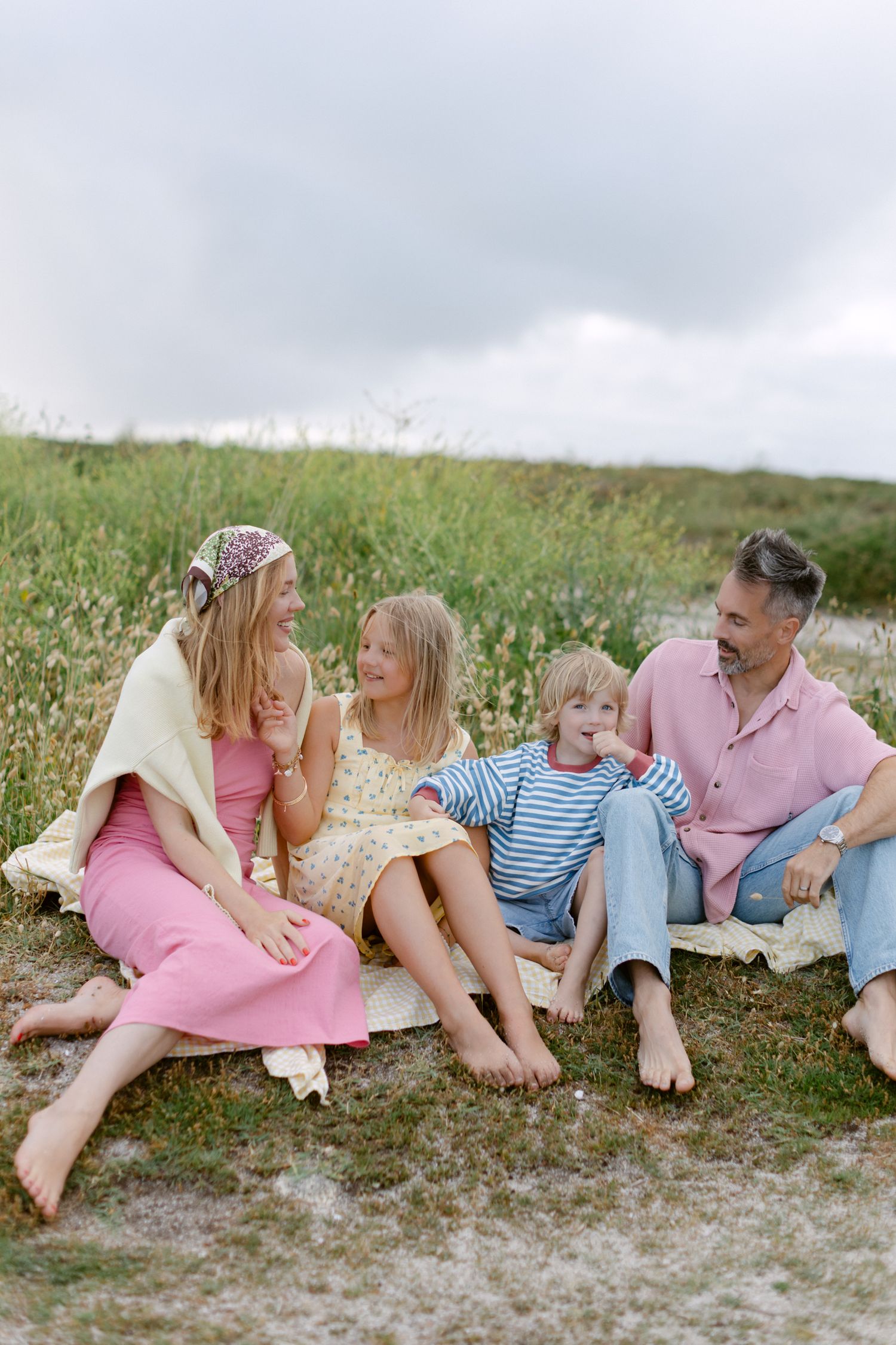 Happy family of five sitting on a blanket in a grassy field, enjoying a casual outdoor picnic together.