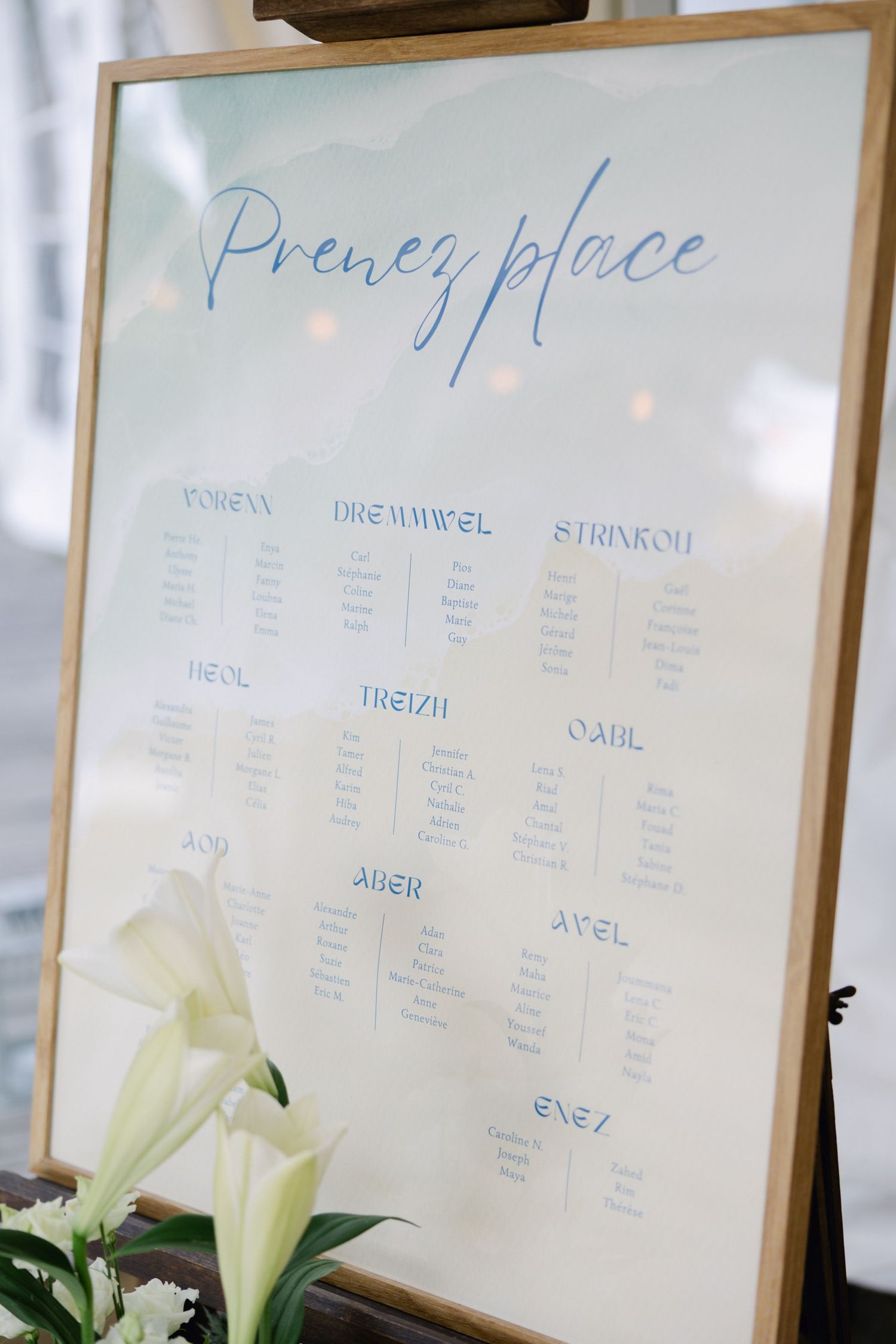 Elegant French wedding seating chart in blue script on white board reading Prenez Place in wooden frame with white lilies.