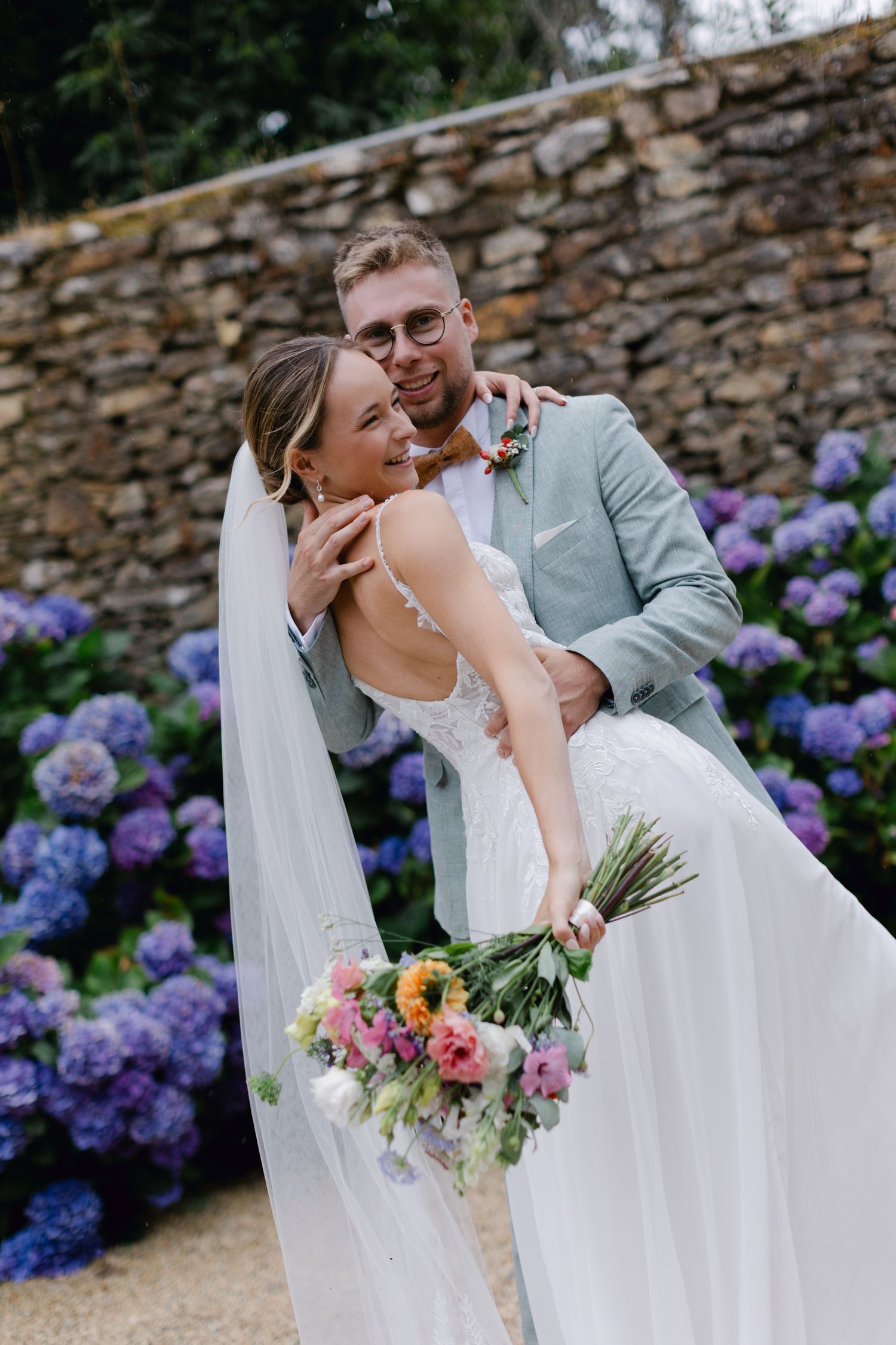 Le marié en costume de sauge soulève une mariée souriante en robe en dentelle avec un bouquet coloré devant les hortensias violets.
