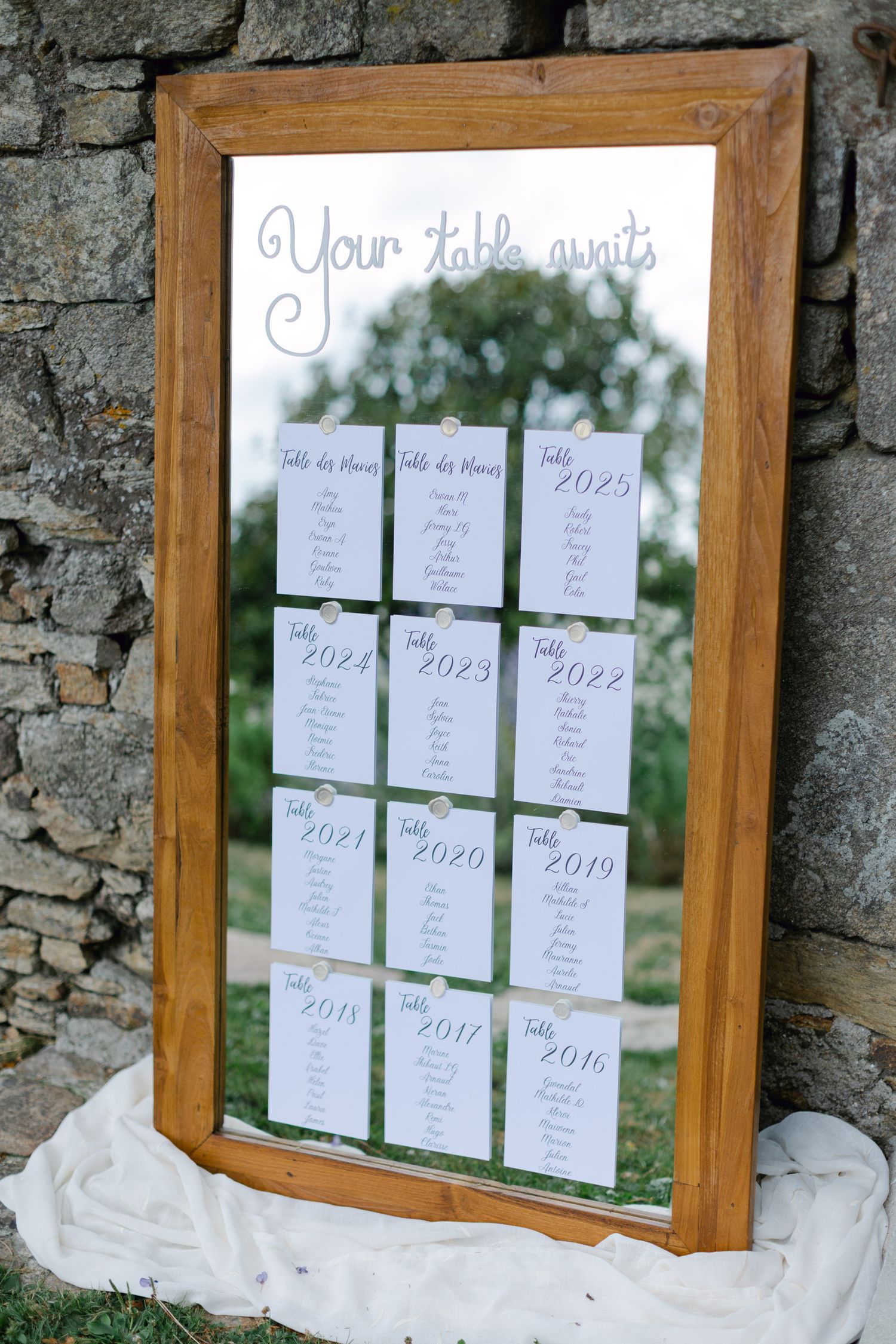 Wedding seating table displayed on a rustic wooden mirror with the script Your Table Awaits against a stone wall.