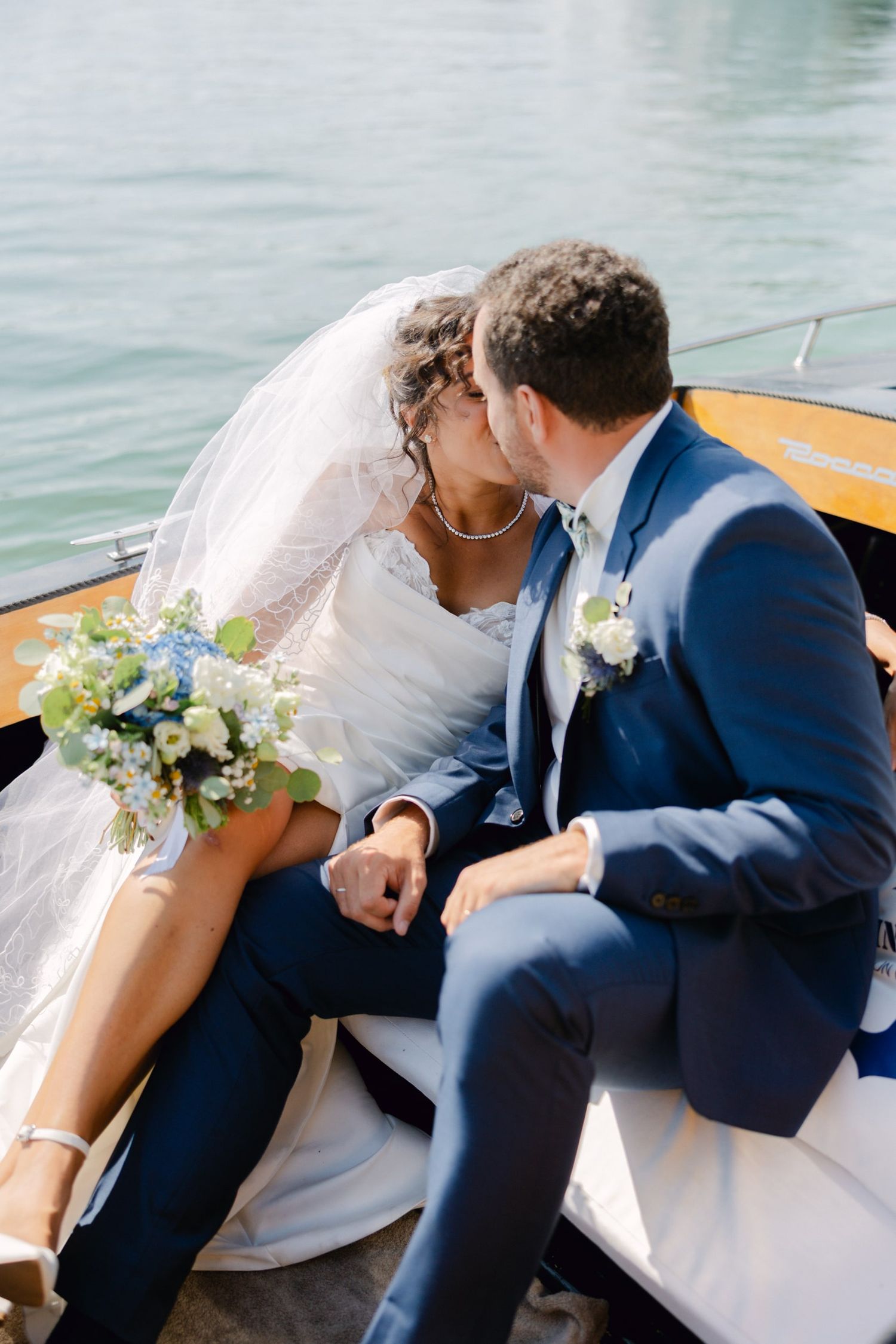 Bride and groom kissing on a boat holding blue floral bouquet on their wedding day.