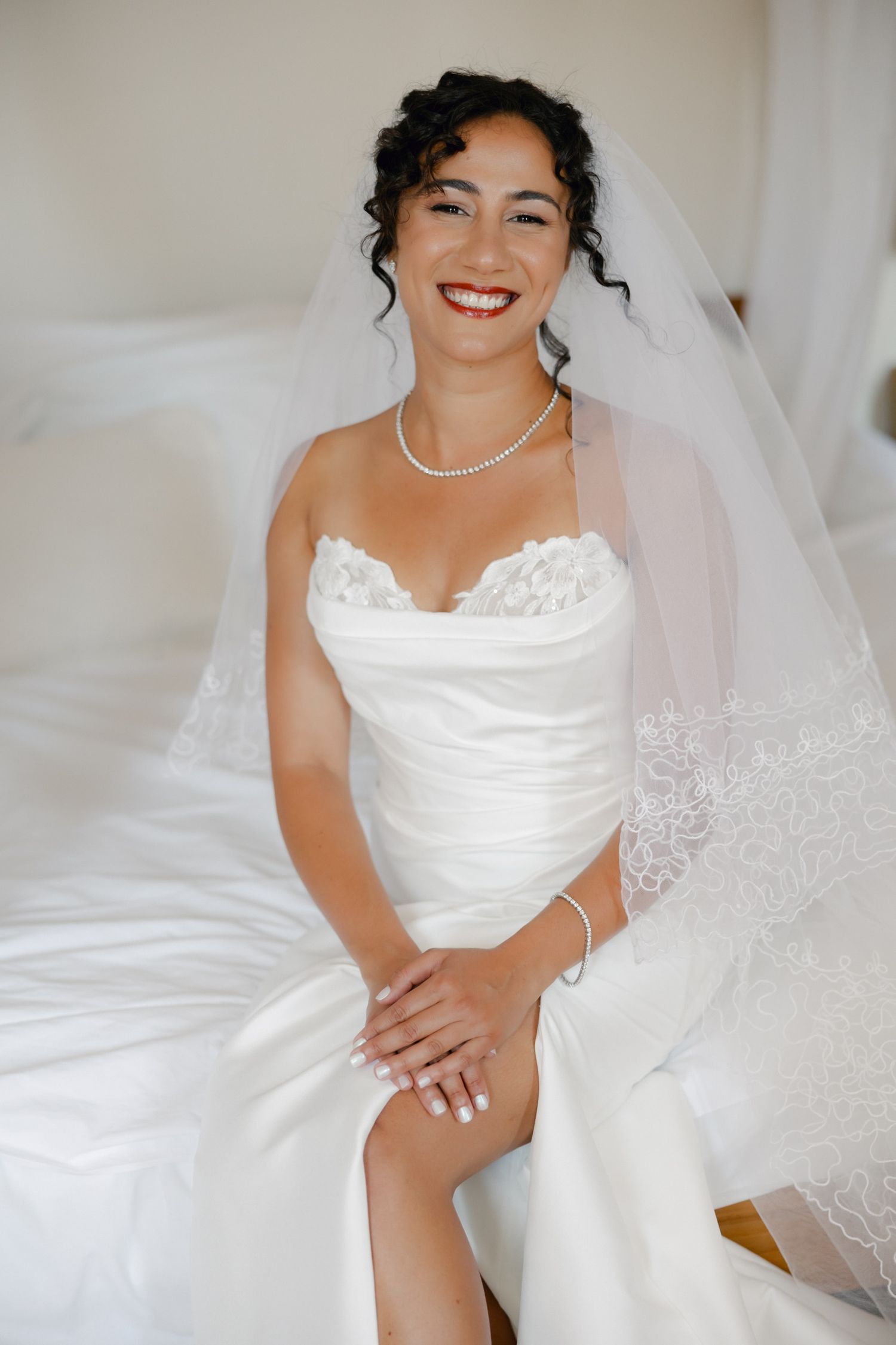 Smiling bride in white strapless gown with lace veil and pearl jewelry sits on bed on her wedding day.