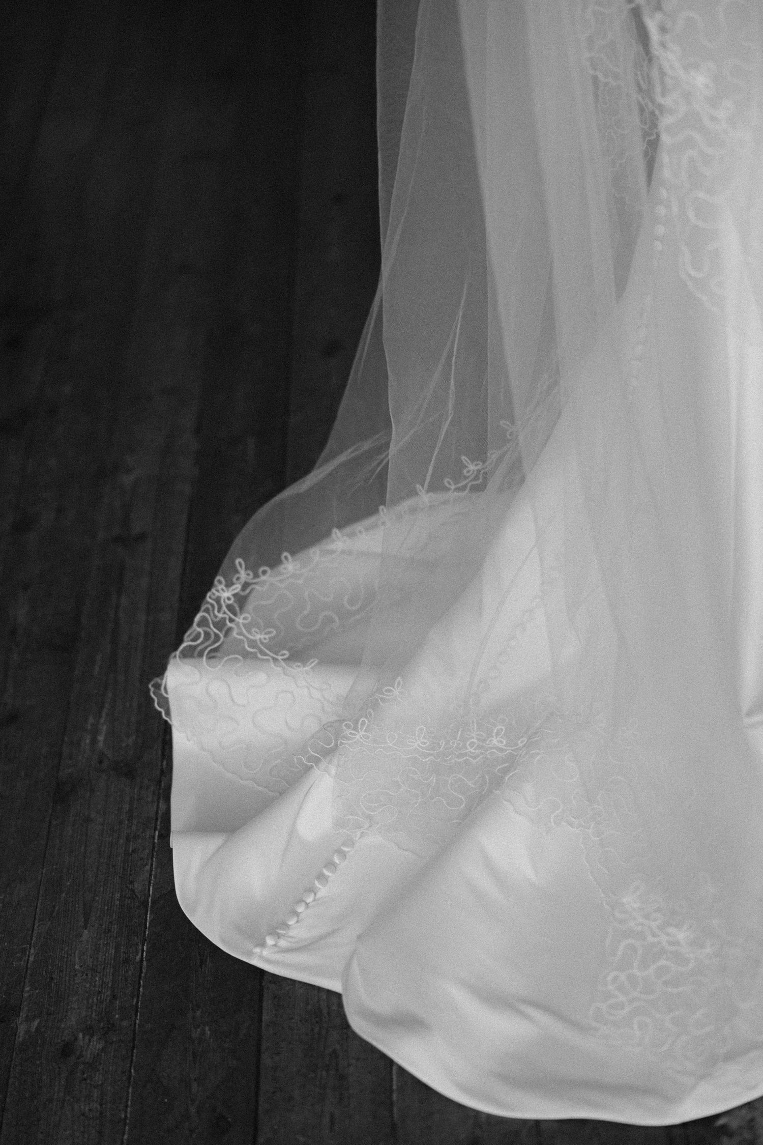 Black and white photo of a bridal gown train and veil with delicate lace detailing on a dark hardwood floor.