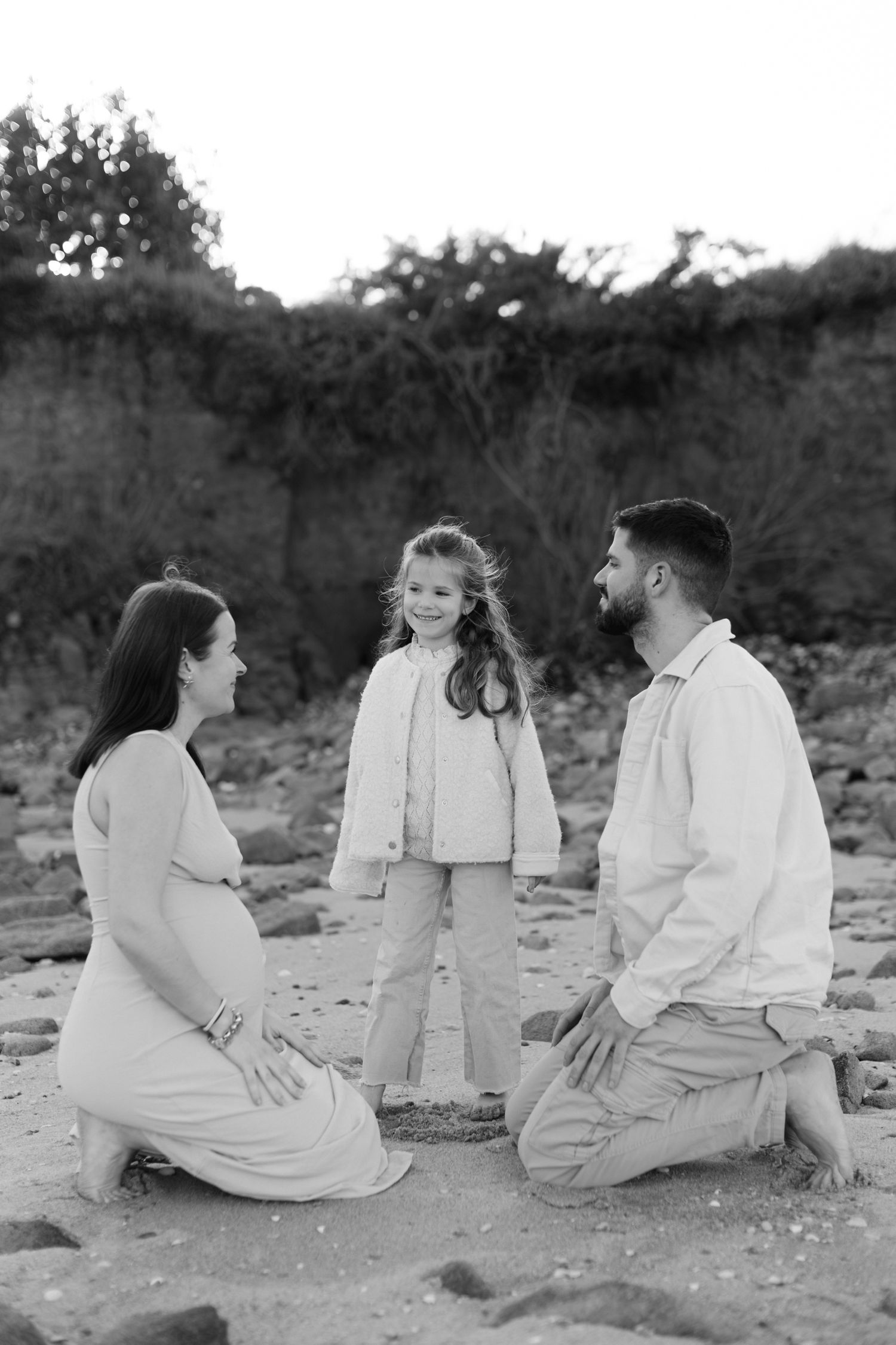 Photo en noir et blanc d'une femme enceinte et d'un homme agenouillés sur la plage face à une jeune fille souriante entre eux.