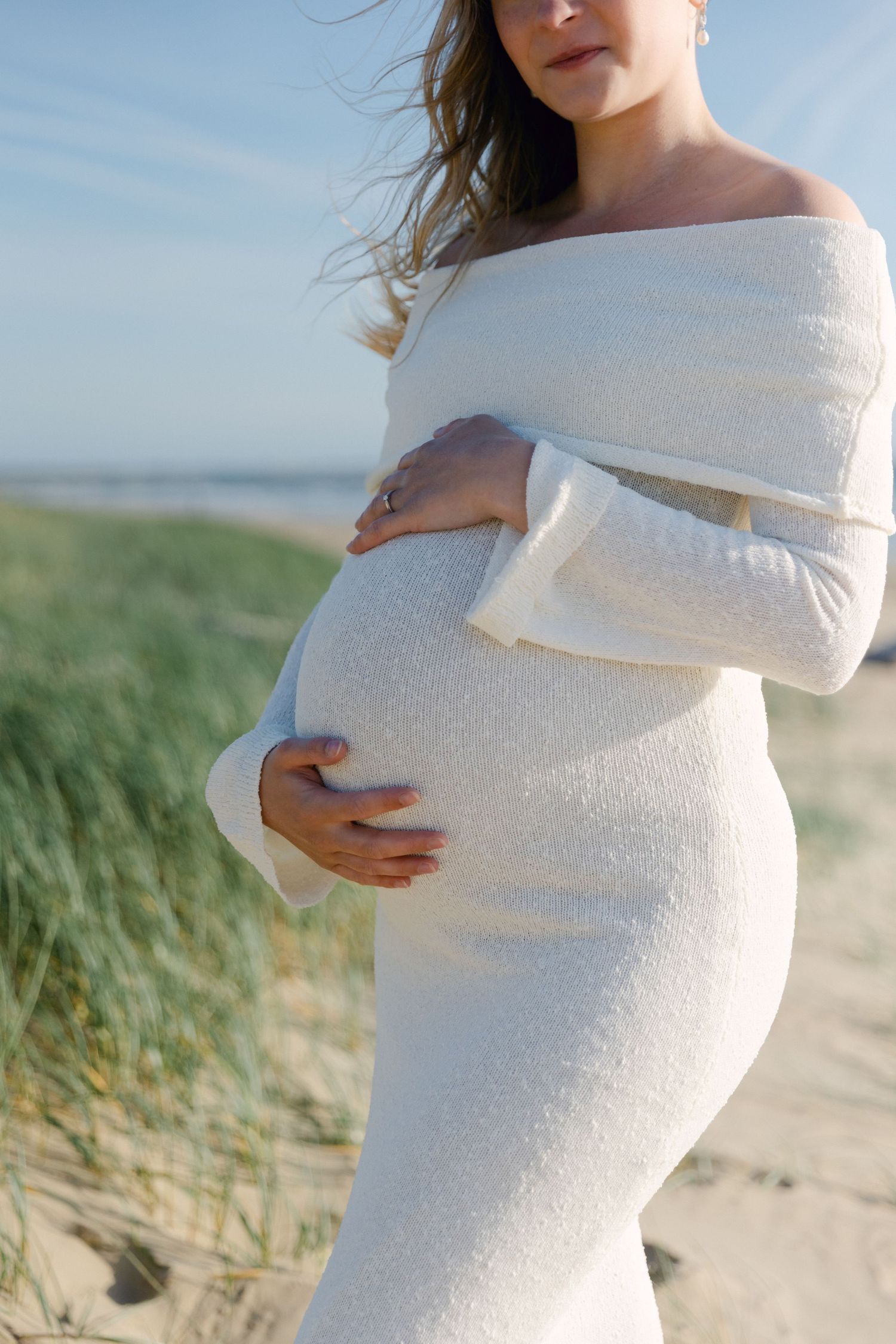 Femme enceinte en robe blanche à épaules dénudées berçant sa bosse de bébé sur une plage de sable avec des dunes.