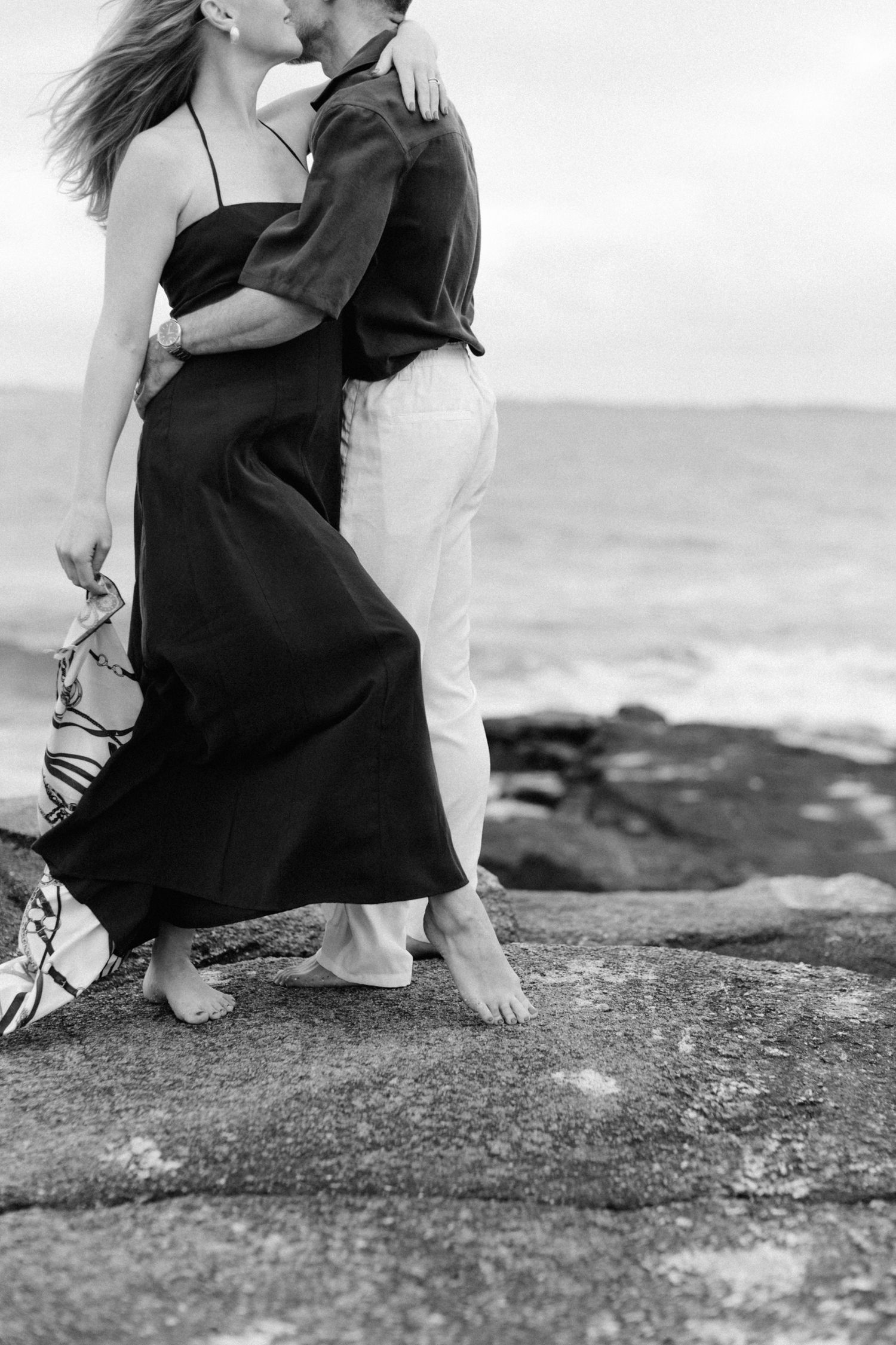 Photo en noir et blanc d'un couple s'embrassant pieds nus sur des rochers côtiers avec les vagues de l'océan en arrière-plan.