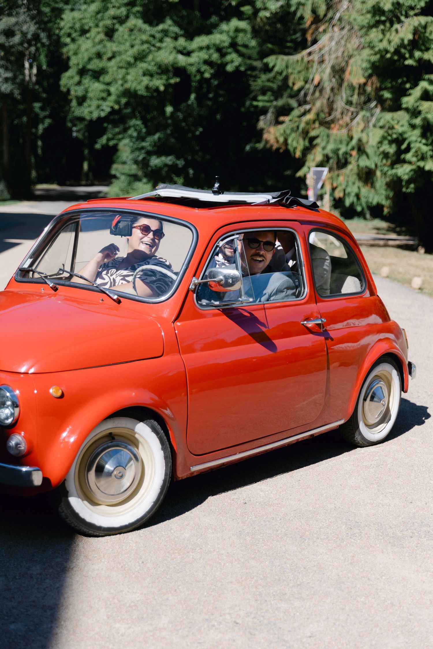 Deux personnes conduisant une Fiat 500 classique rouge sur une route ensoleillée bordée d'arbres.