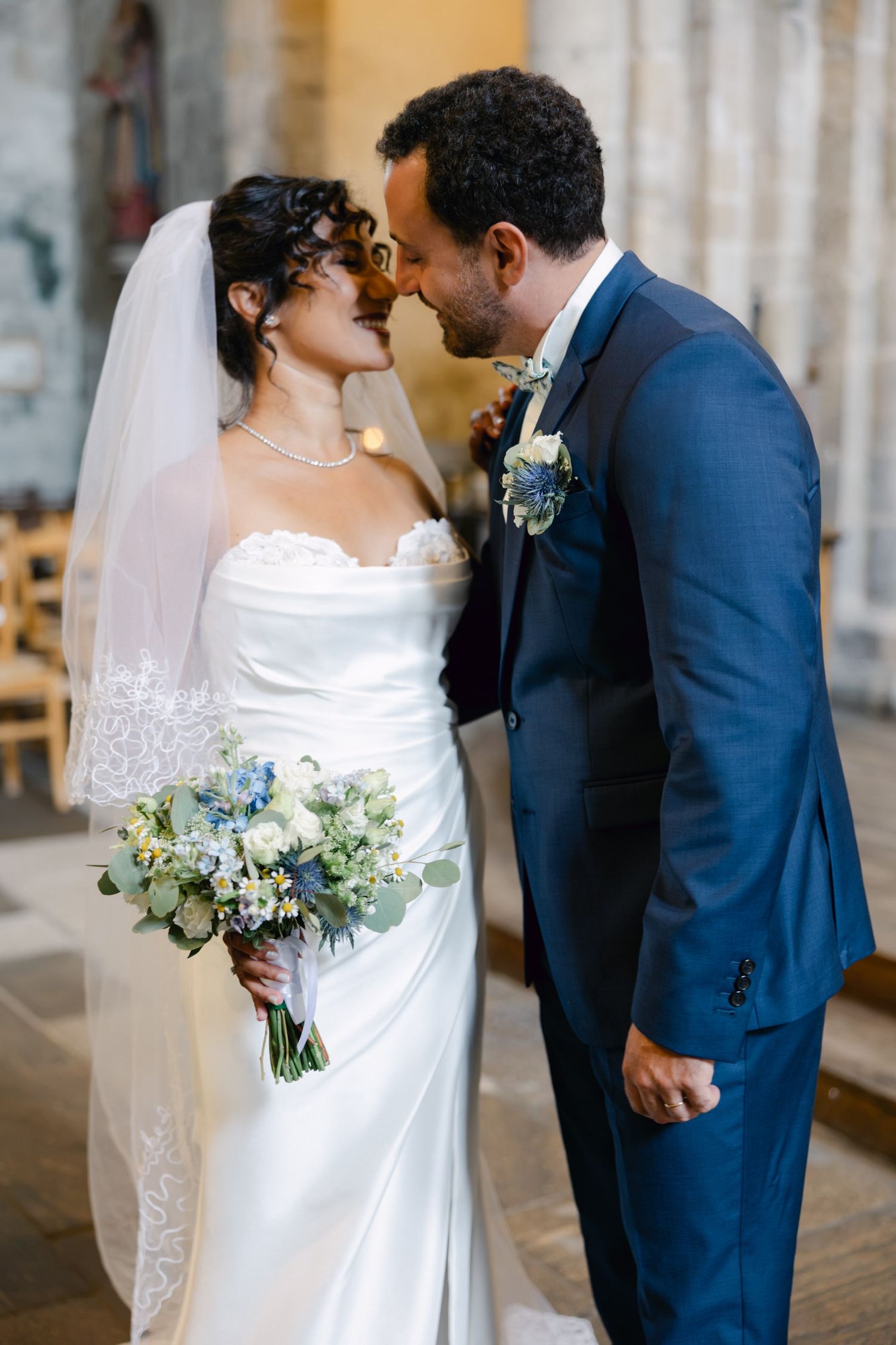 Bride and groom share an intimate moment, nearly kissing, as she holds a blue and white bouquet in a church.
