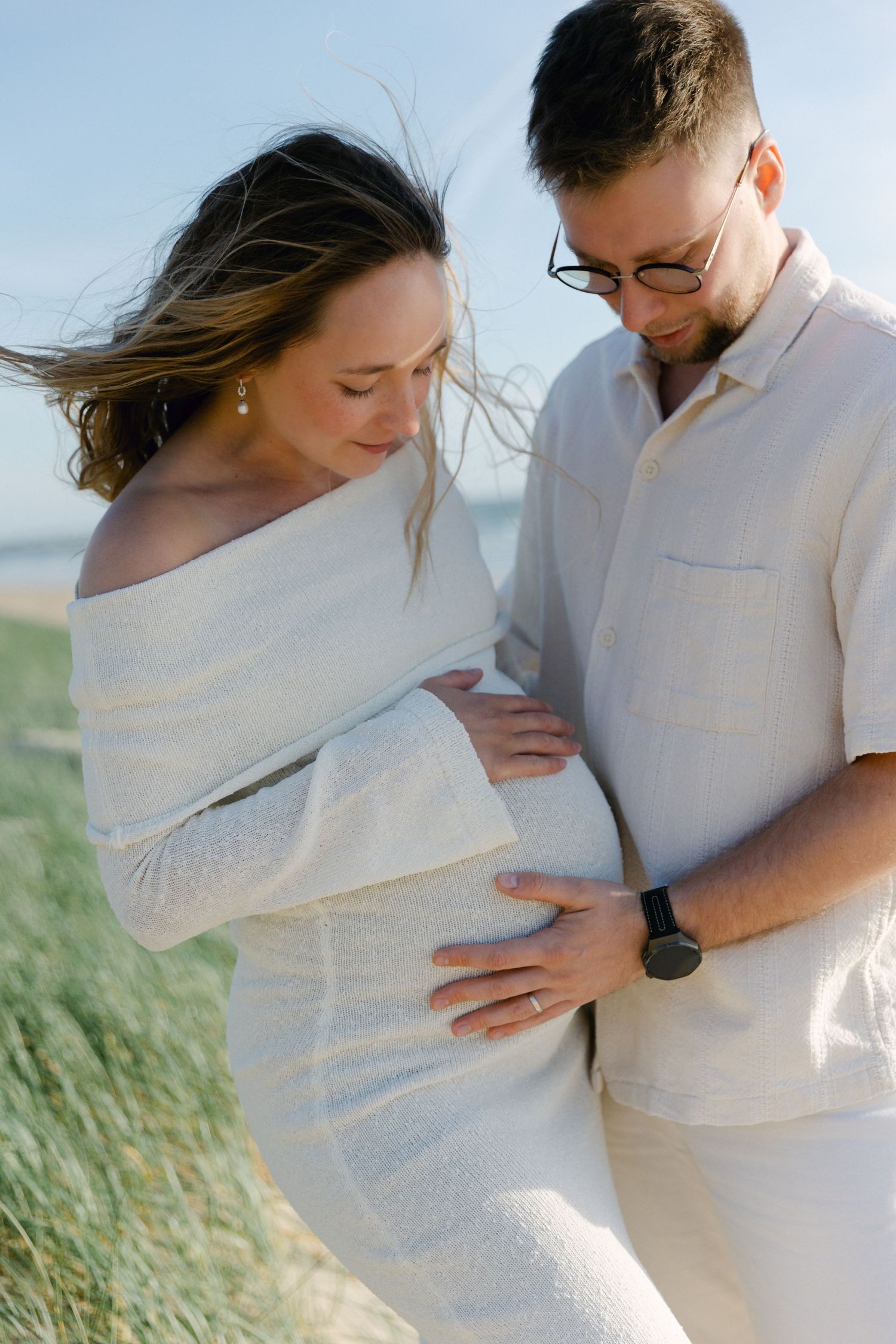 Couple en tenue blanche regardant tendrement le ventre de femme enceinte ensemble sur une plage venteuse.