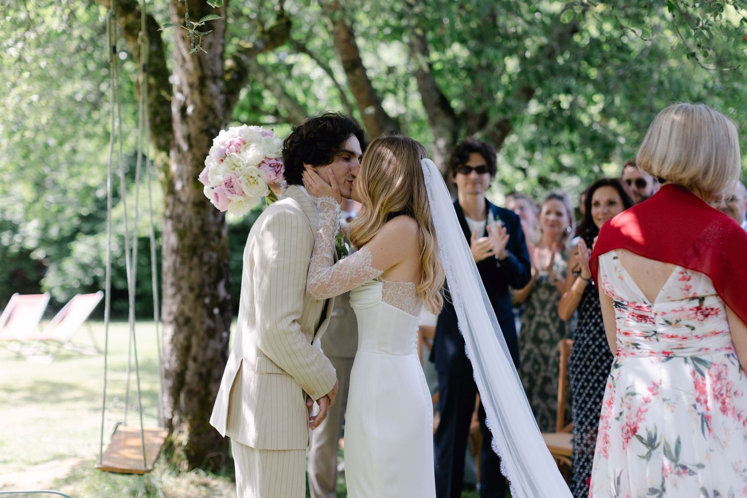 La mariée et le marié partagent leur premier baiser lors d'une cérémonie de mariage en plein air entourée d'invités et d'arbres verts.