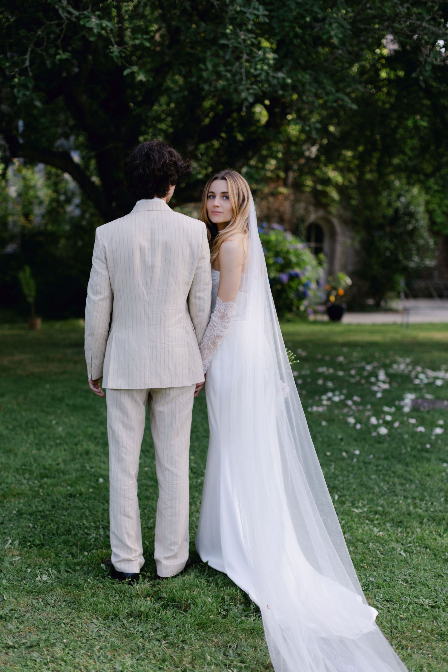 La mariée en robe blanche avec un long voile regarde en arrière tout en se tenant à côté du marié en costume beige dans un cadre de jardin.