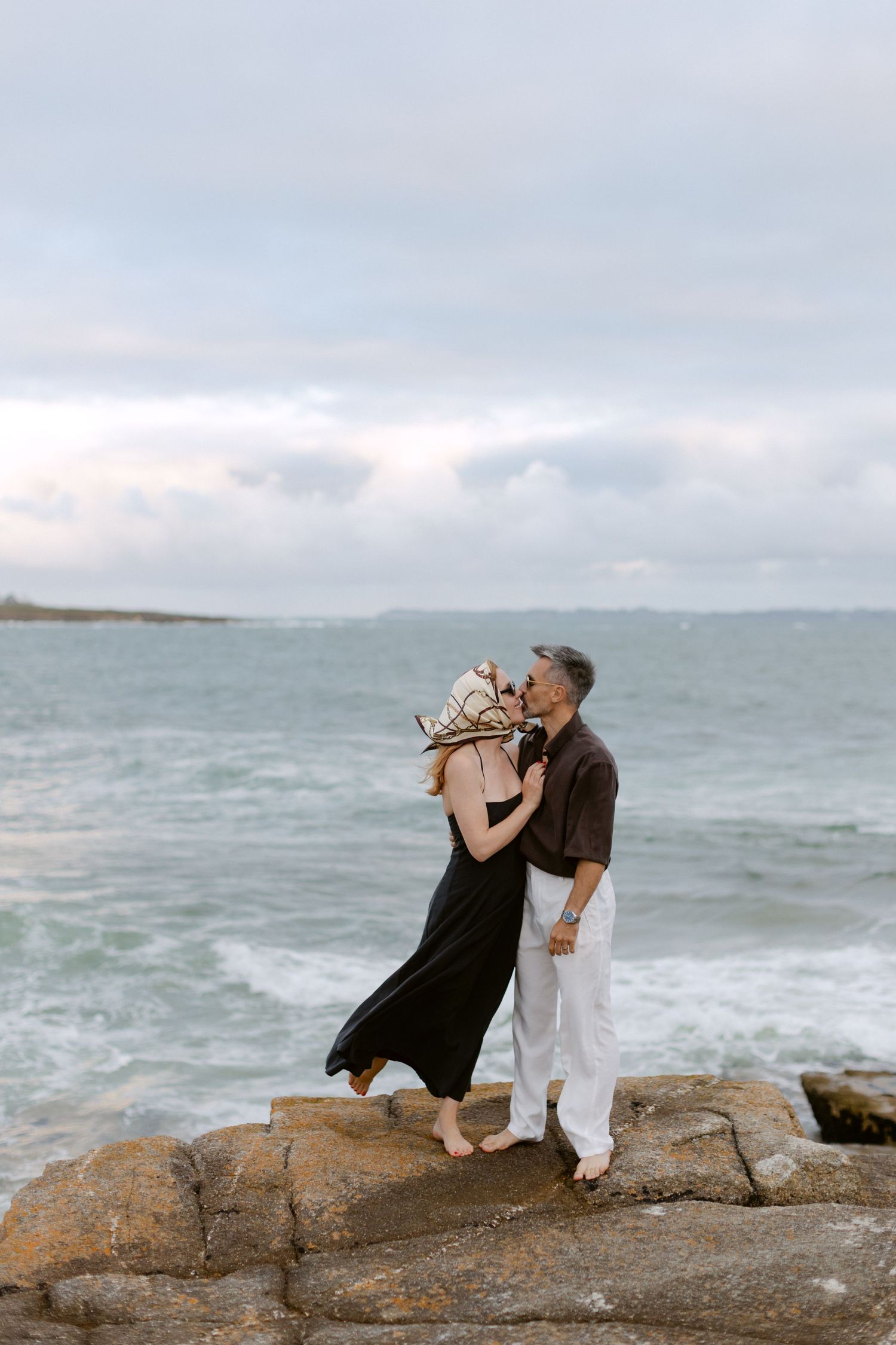 Couple embracing on coastal rocks by the ocean, woman in black dress and floral headscarf, man in white pants.
