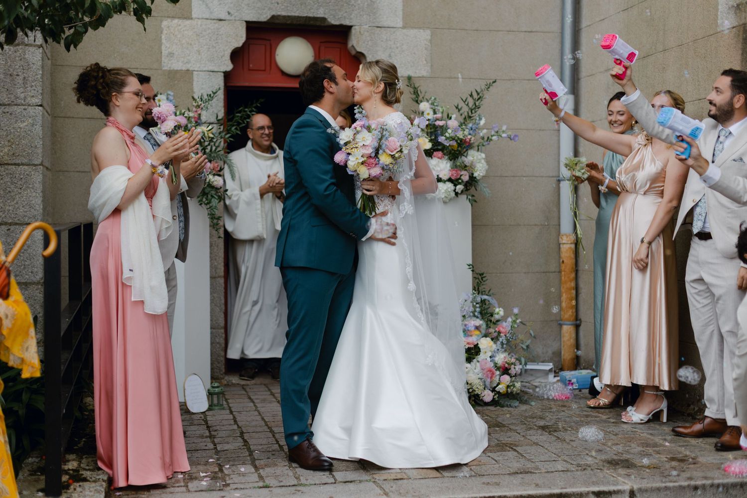 Les jeunes mariés partagent leur premier baiser à l'extérieur de l'entrée de l'église alors que les invités célèbrent avec des confettis et