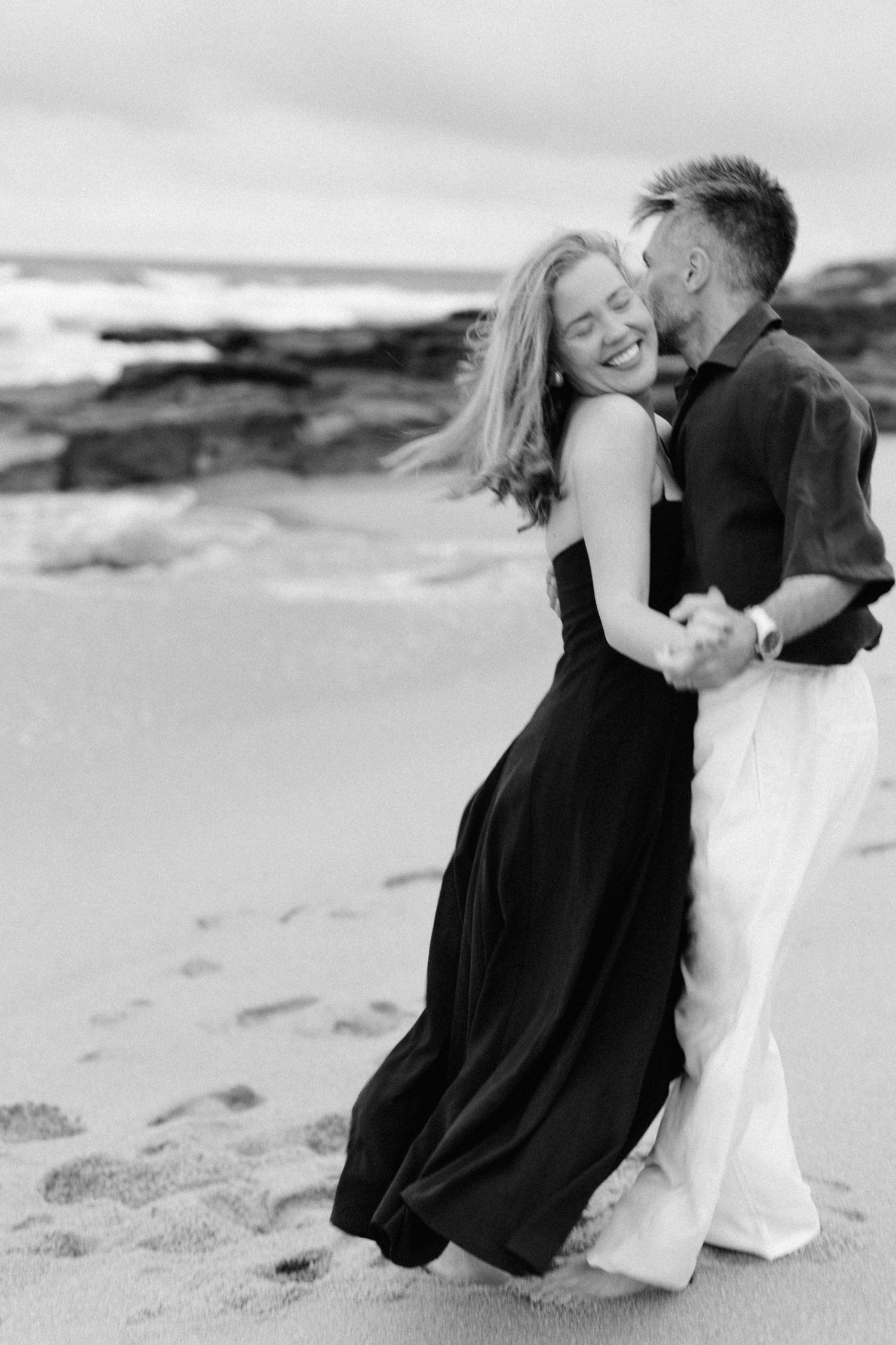 Couple s'embrassant et riant sur une plage de sable en noir et blanc, femme en robe fluide, photo de fiançailles côtières romantiques.