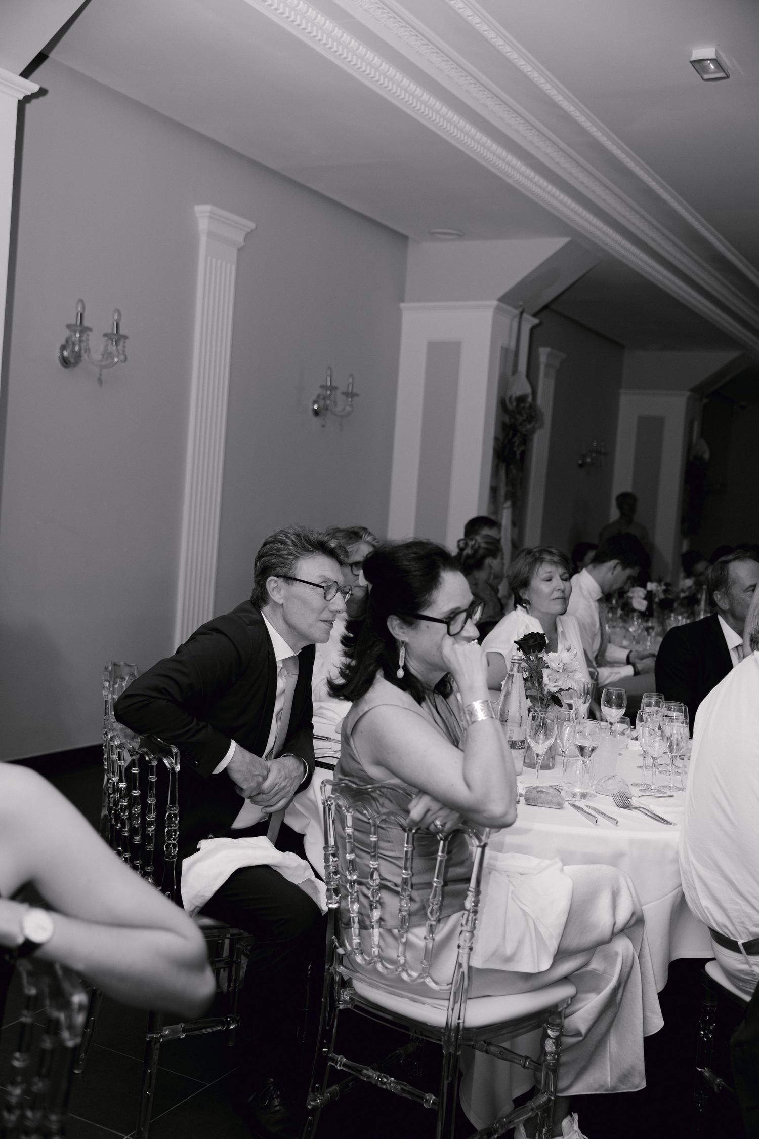 Photo en noir et blanc d'invités de mariage élégamment habillés assis à une table de réception dans un lieu formel.