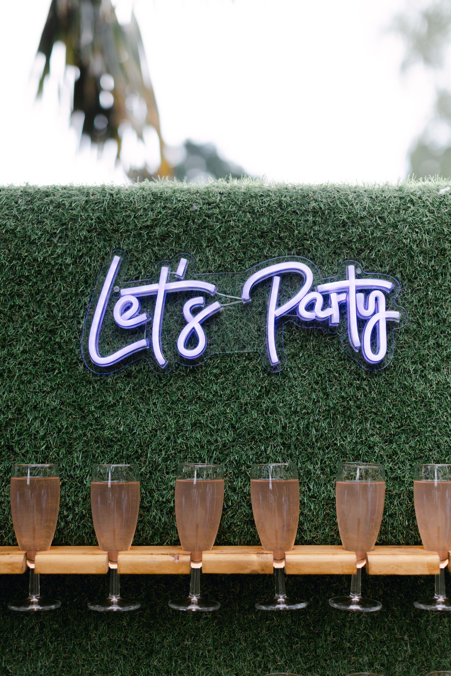 Panneau Neon Let's Party sur un mur de haie verte au-dessus d'une rangée de flûtes à champagne au bar de réception de mariage.