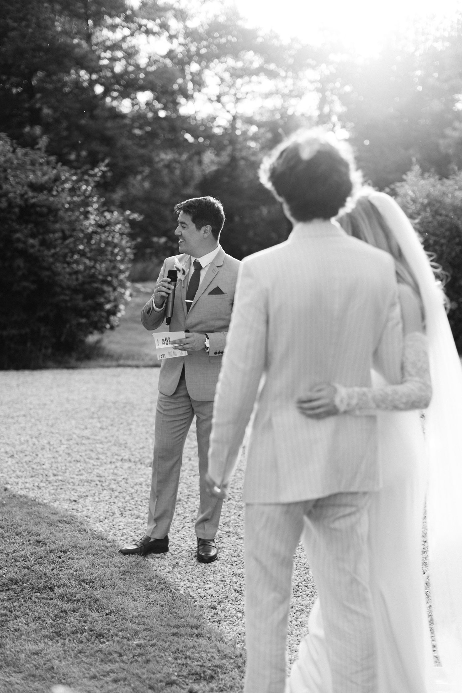 Photo en noir et blanc d'un marié parlant lors d'une cérémonie de mariage en plein air au soleil.