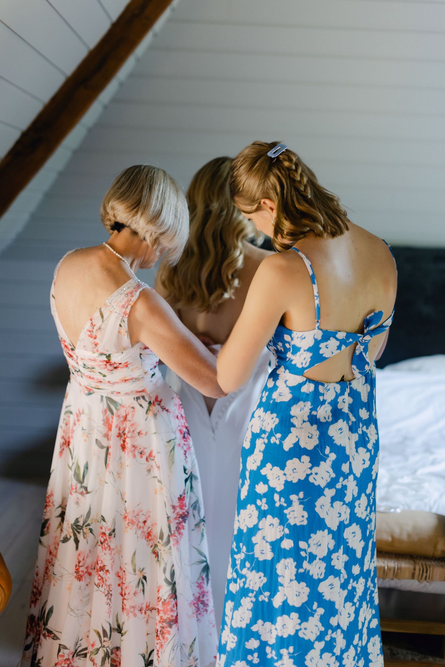 Deux demoiselles d'honneur en robes à fleurs aident une mariée à attacher sa robe de mariée blanche dans une pièce rustique.