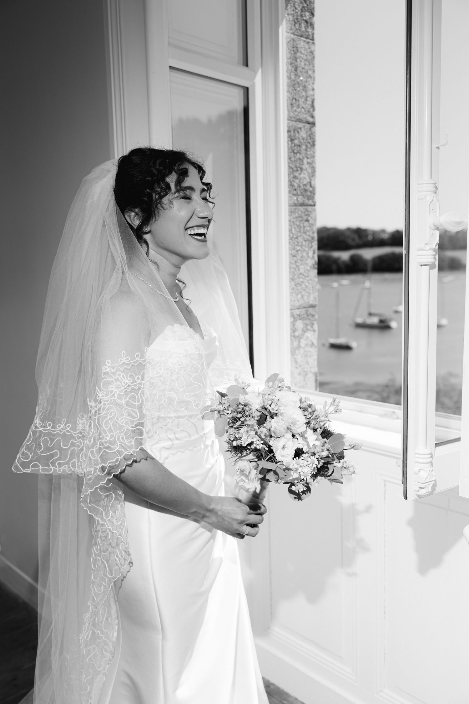 Smiling bride in strapless gown and lace veil holds bouquet by window in black and white wedding photo.