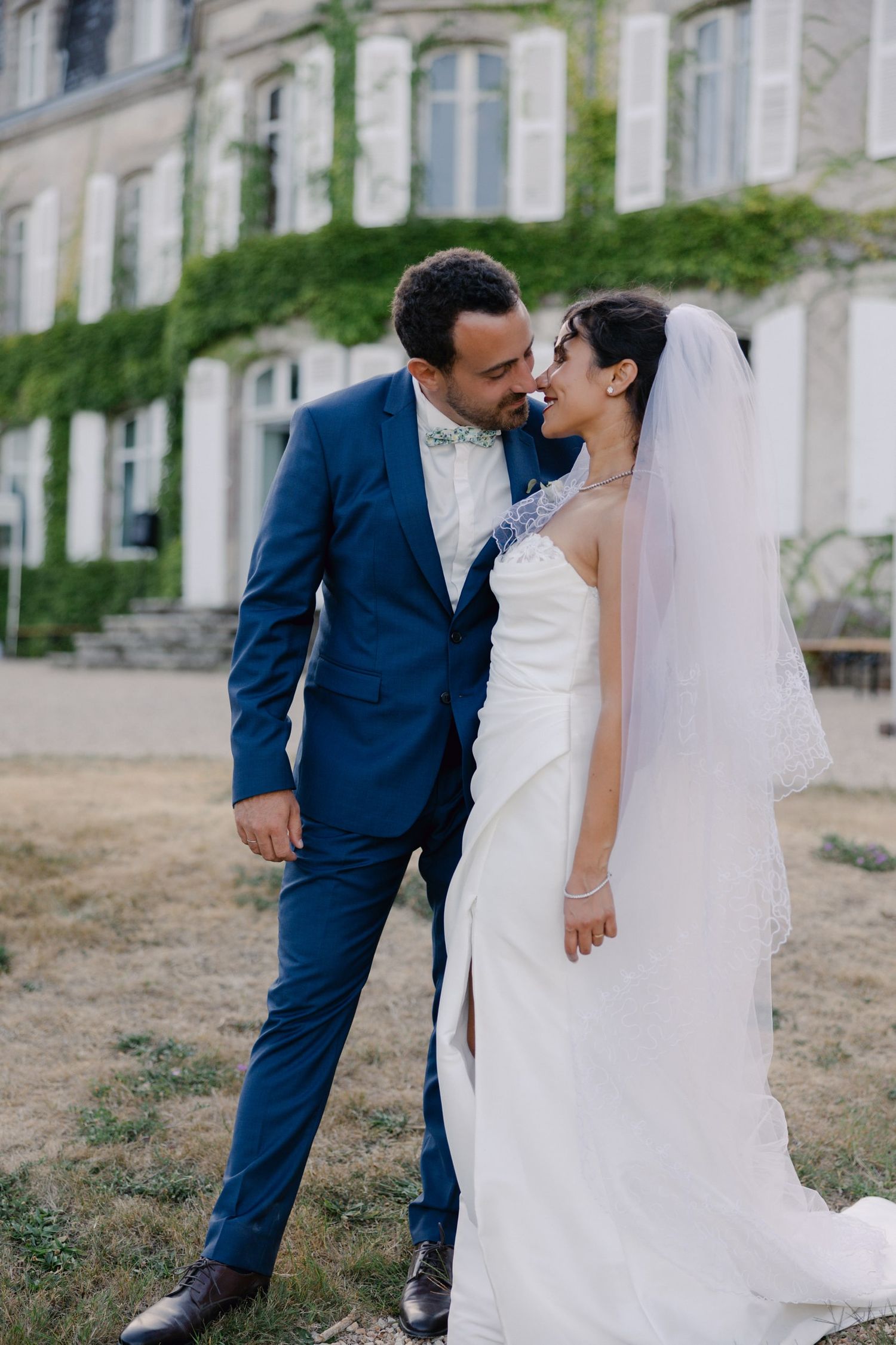 A bride and groom share a romantic moment outside an ivy-covered French chateau, dressed in a white gown and blue suit.
