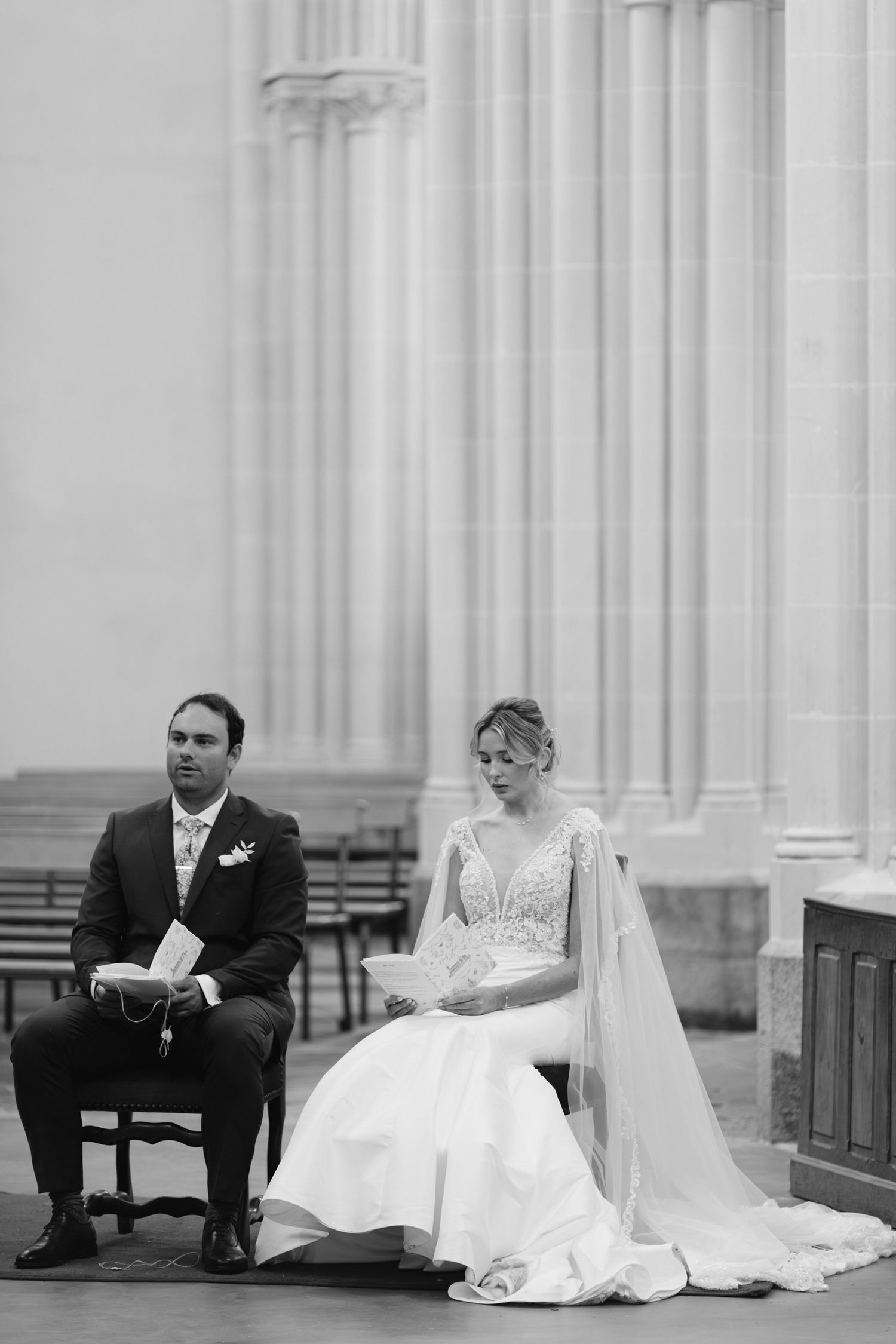 Photo en noir et blanc de la mariée et du marié assis lors de la cérémonie de mariage à l'église à côté de colonnes de pierre.