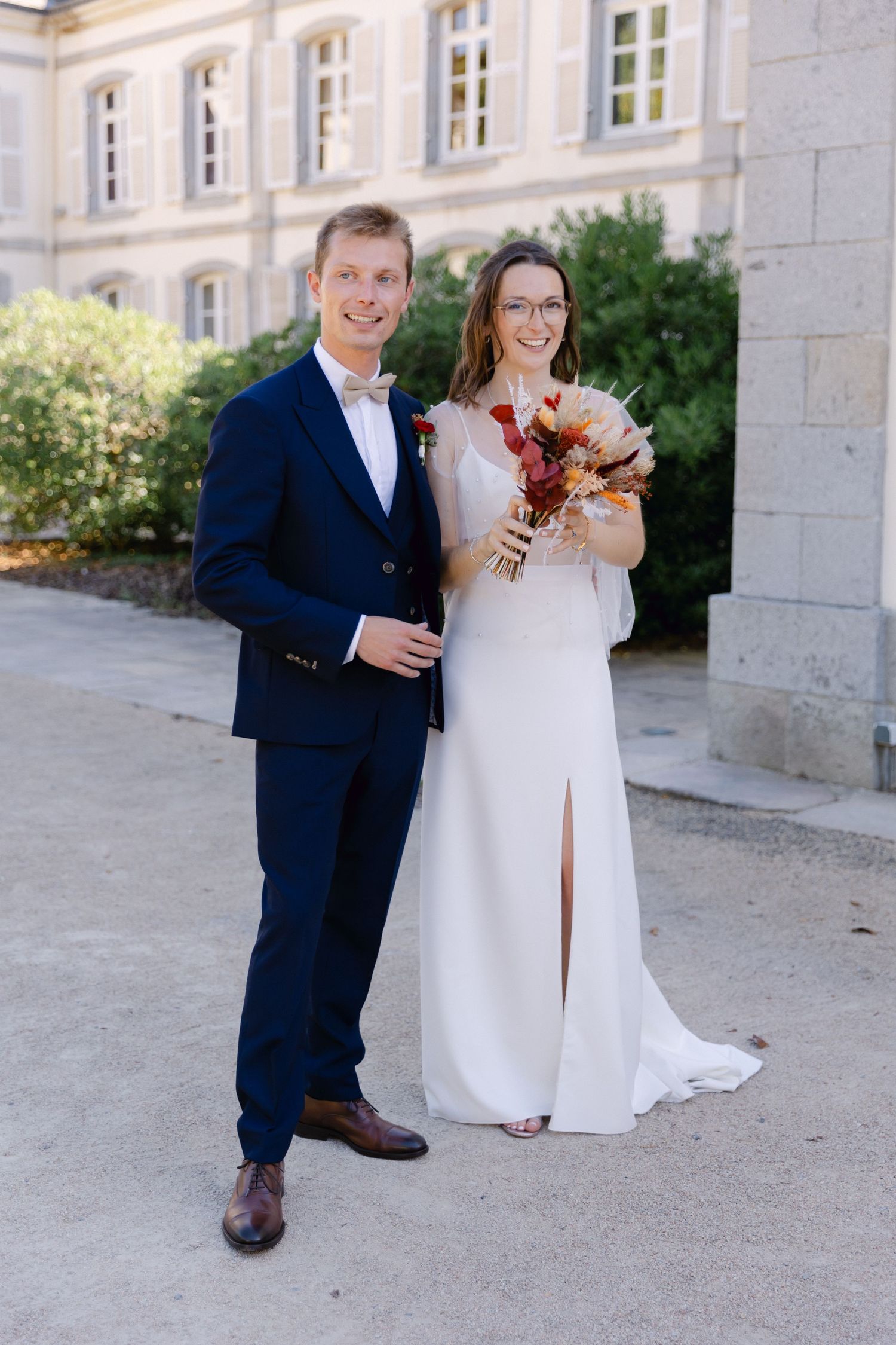 La mariée et le marié posant à l'extérieur en tenue de mariage, elle tient un bouquet de fleurs séchées, une toile de fond élégante.