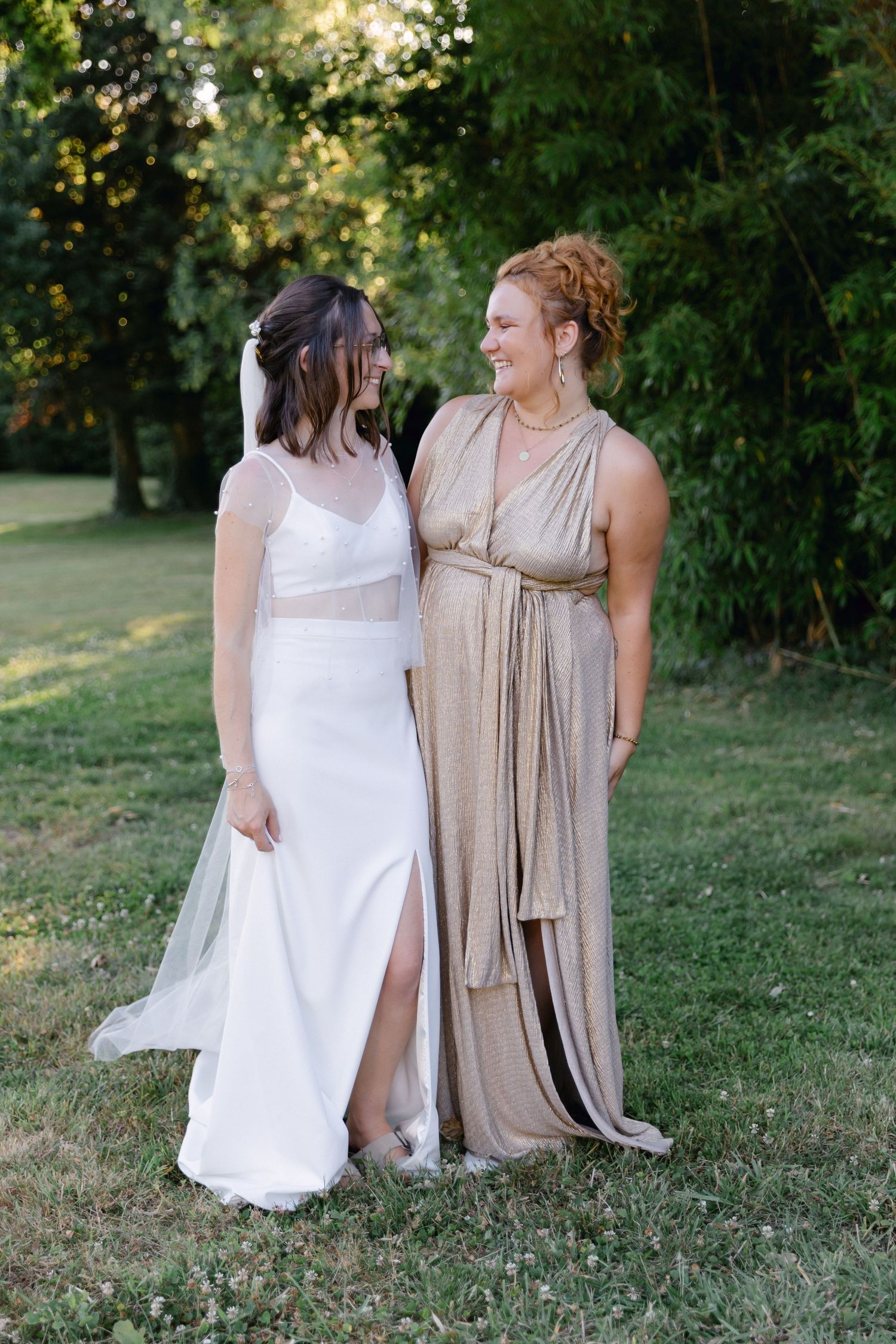 Mariée en robe blanche avec voile se tient à côté de la demoiselle d'honneur en robe portefeuille dorée, souriant à l'extérieur.