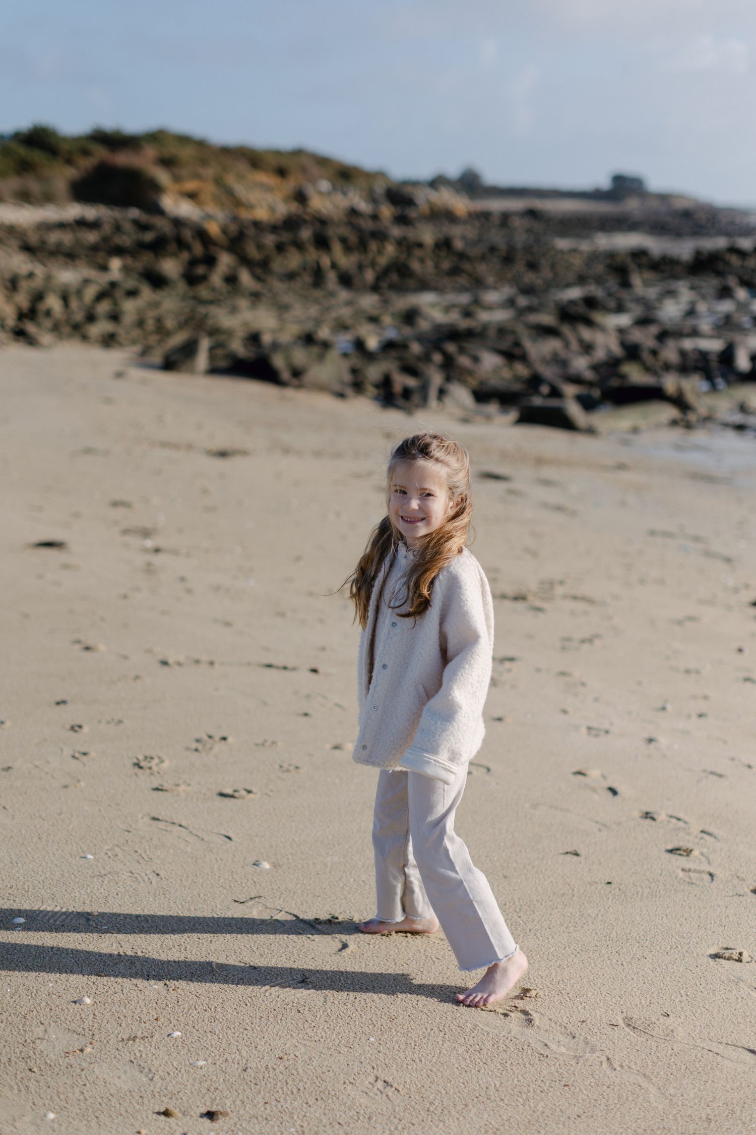 Jeune fille heureuse en tenue crème marchant pieds nus sur une plage de sable avec un littoral rocheux derrière elle.