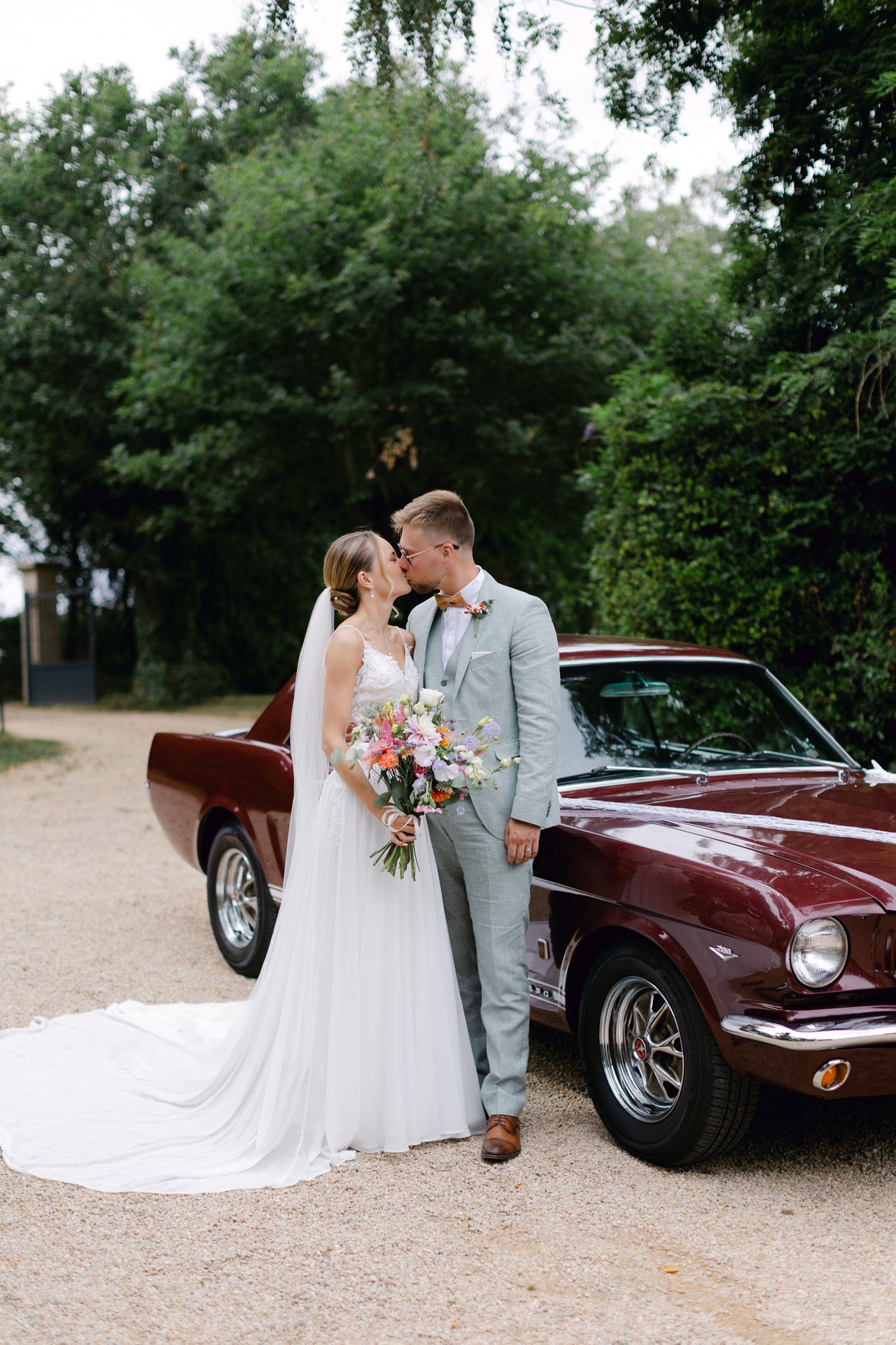 La mariée et le marié posent romantiquement à côté d'une Ford Mustang bordeaux classique sur l'allée de gravier.