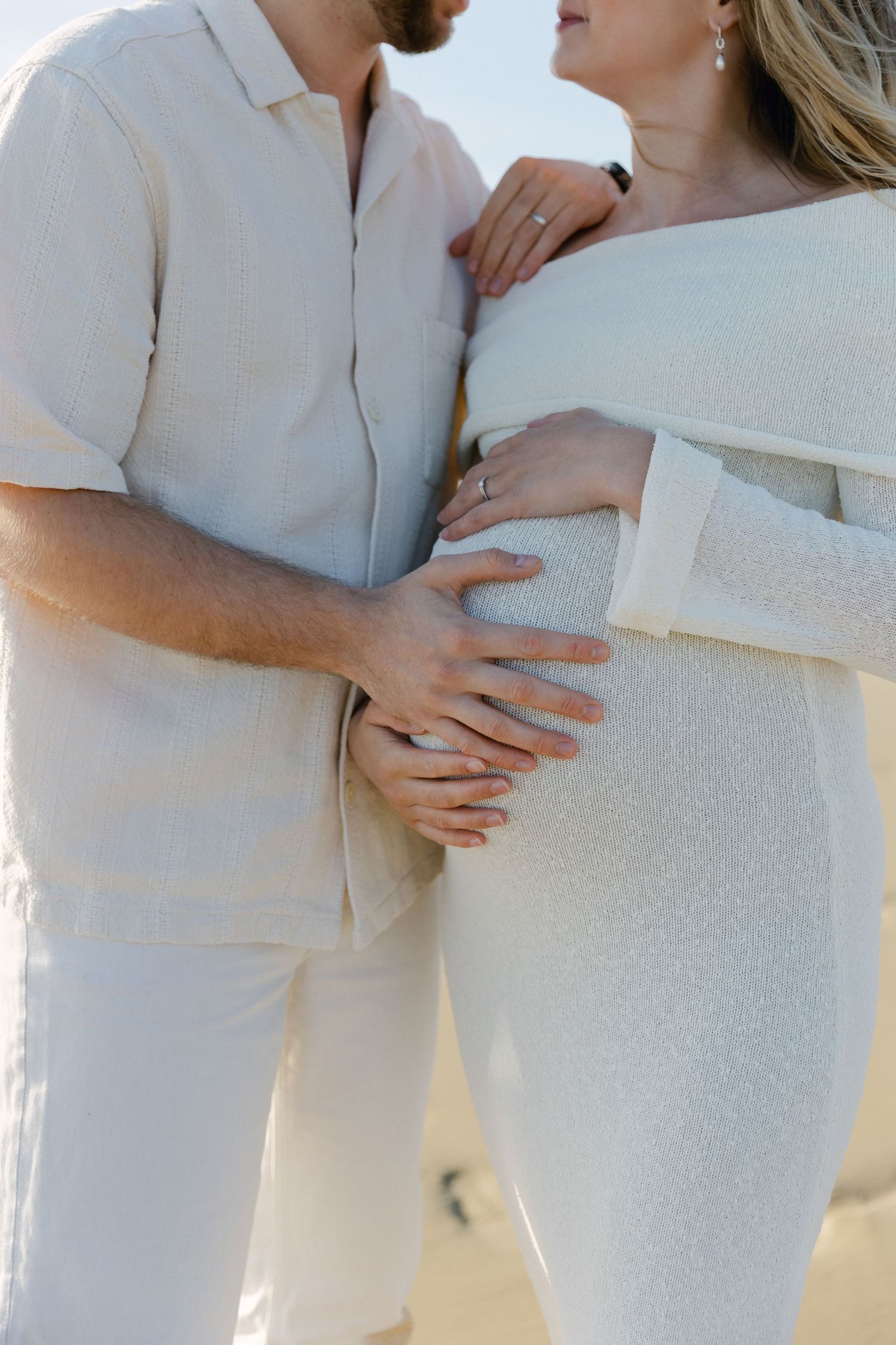 Gros plan d'un couple en tenue blanche tenant le ventre de femme enceinte ensemble sur une plage ensoleillée.