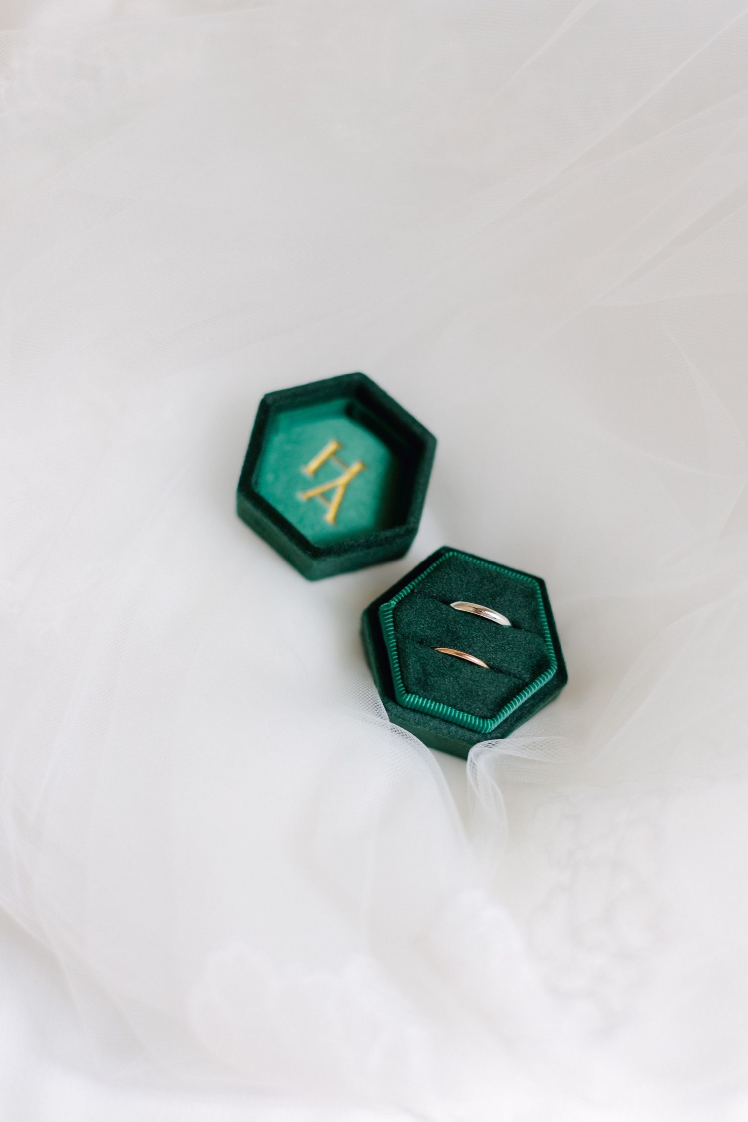 Alliances en or exposées dans une boîte à bagues en velours vert hexagonal ouverte avec des initiales de monogramme en or sur fond blanc.