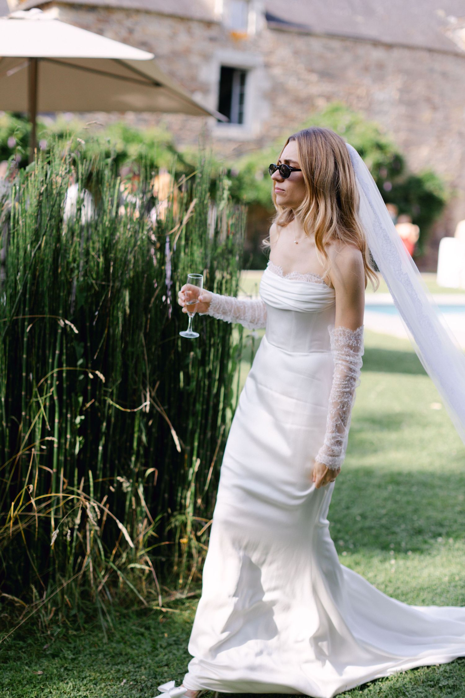 Mariée dans une robe blanche sans bretelles avec de longs gants en dentelle et un voile tenant du champagne près de hautes herbes.