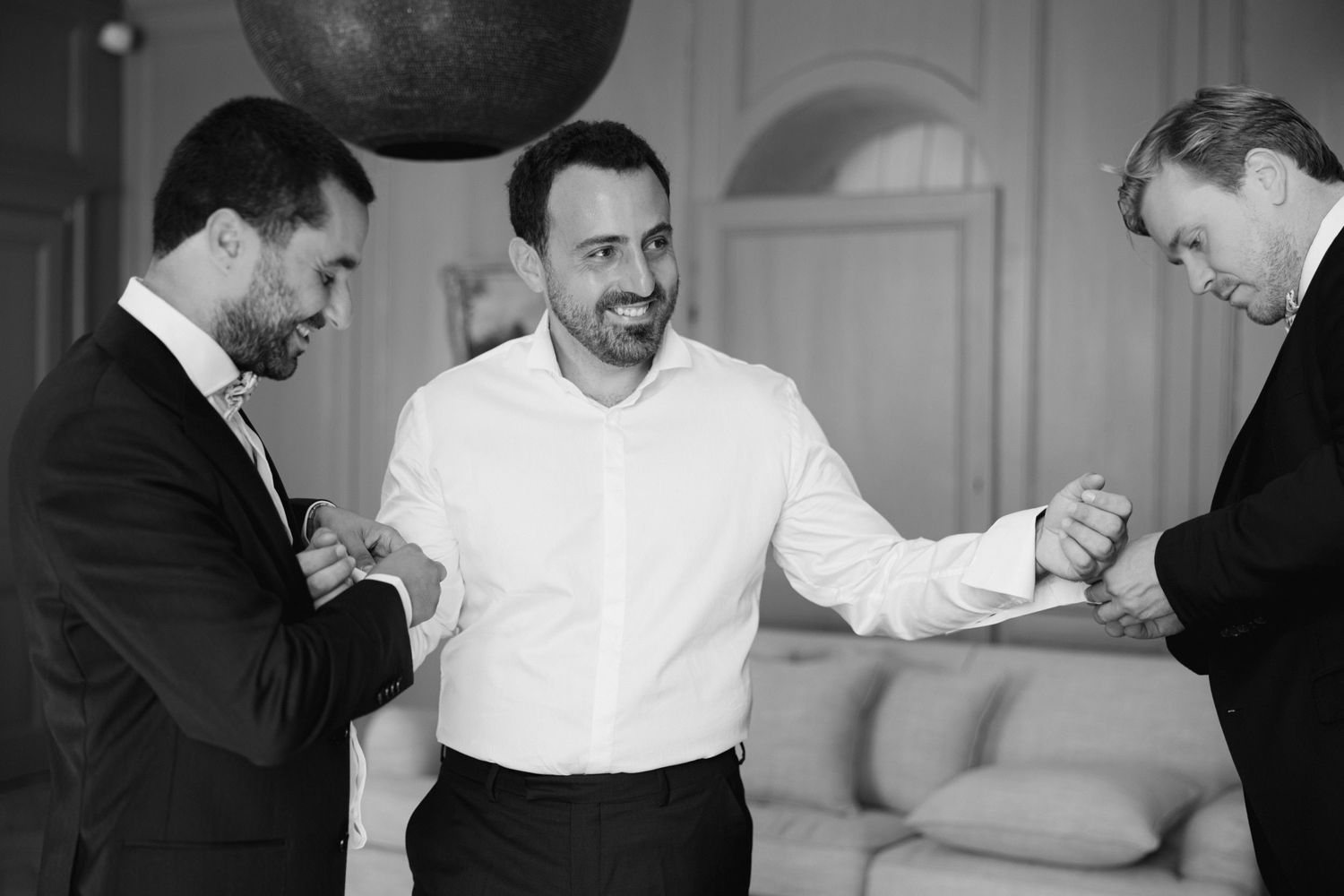 Groom smiling in white dress shirt assisted by two men in suits during wedding preparation in elegant room.