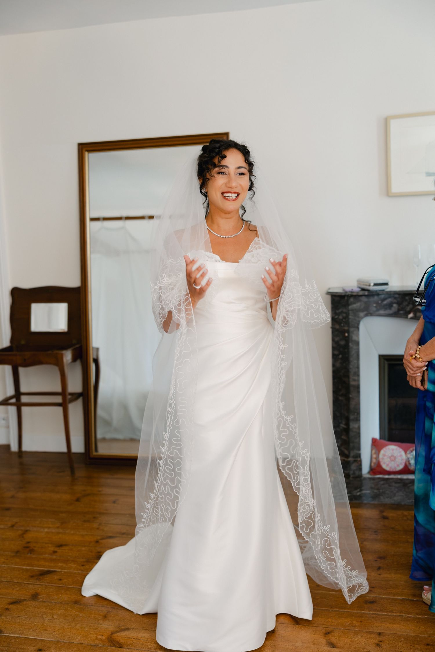 Smiling bride in white gown and lace veil poses indoors on hardwood floors near a fireplace.