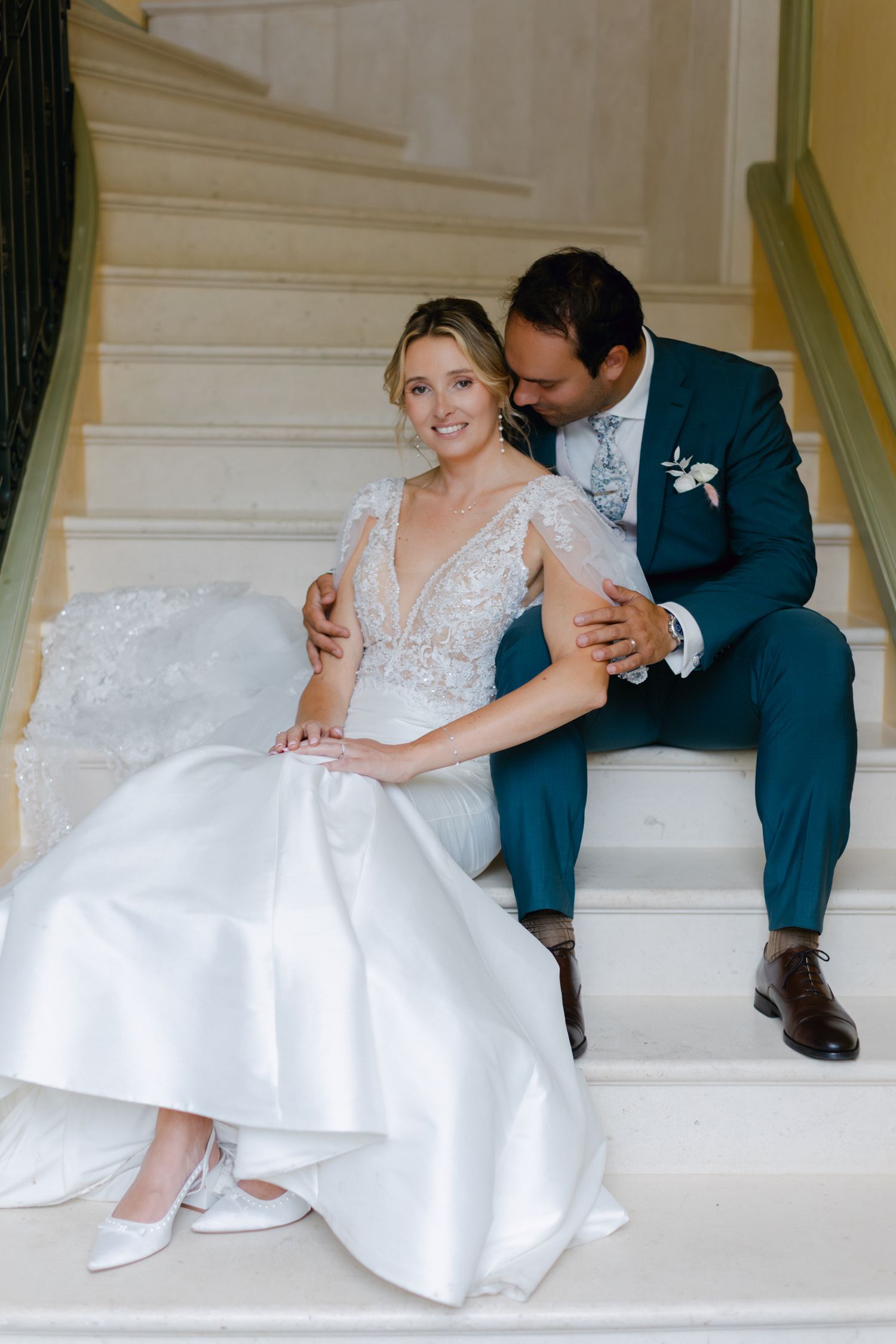 Mariée et marié assis sur un escalier élégant, marié en costume sarcelle embrassant la mariée en robe de mariée en dentelle.