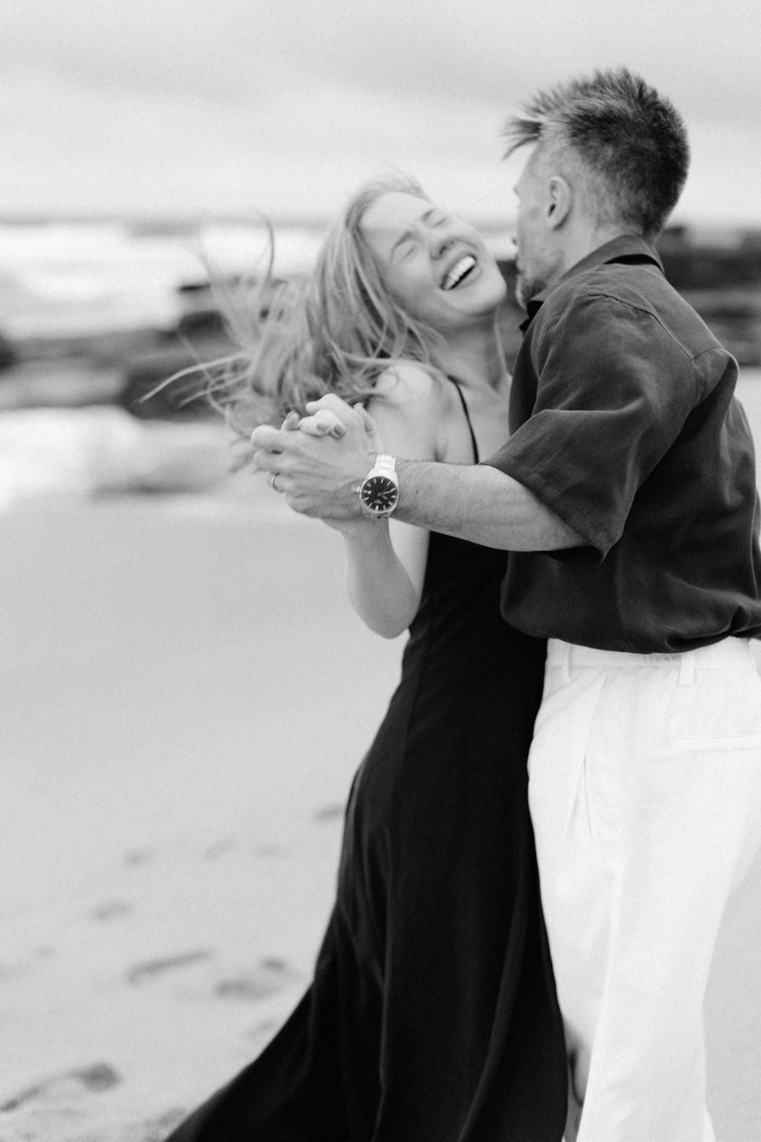 A joyful couple dancing and laughing together on a beach, captured in black and white photography.