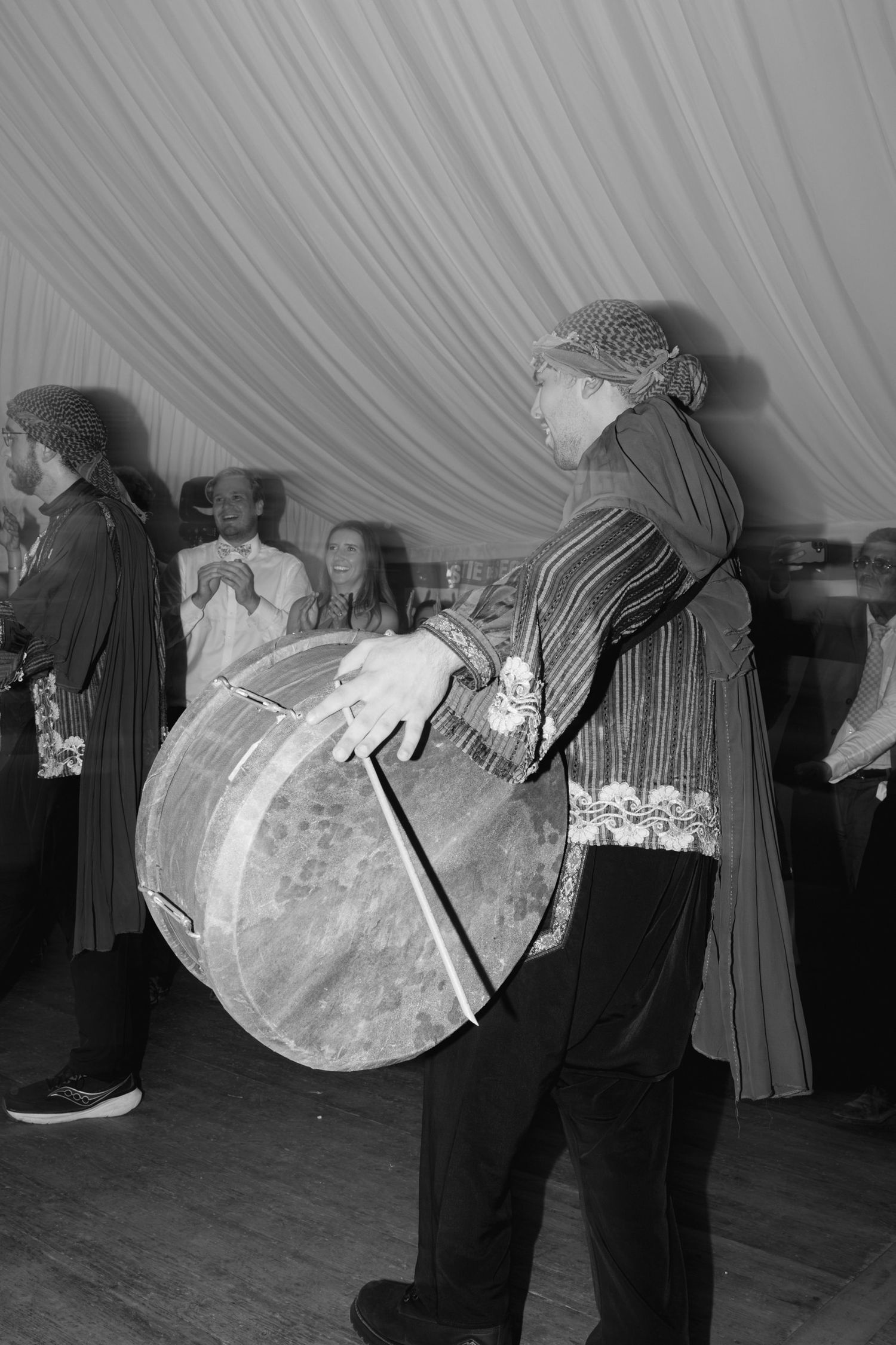 Black and white photo of a musician playing a large drum at a traditional cultural ceremony indoors.