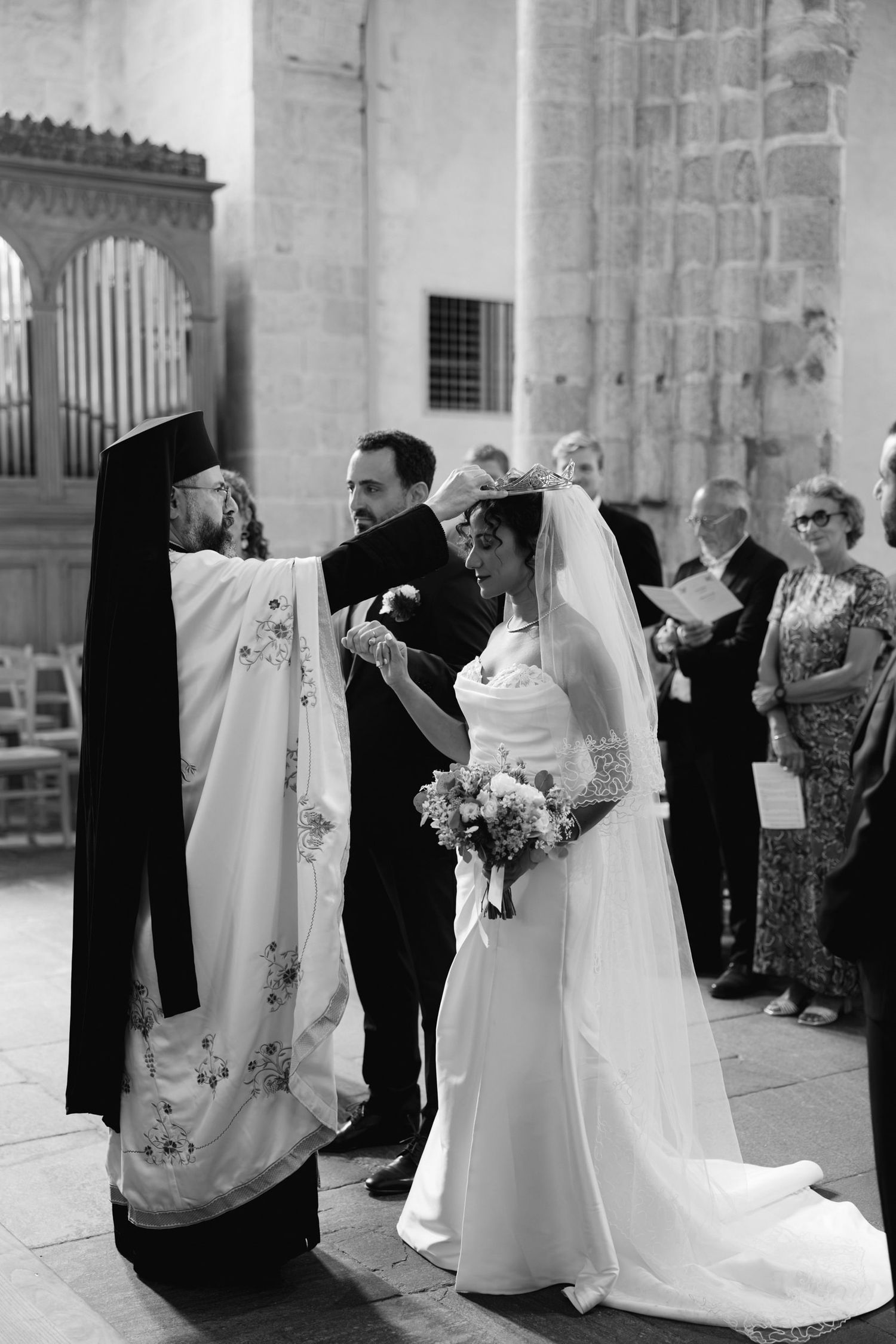 Black and white photo of a bride holding flowers during a church wedding ceremony with a priest and guests.