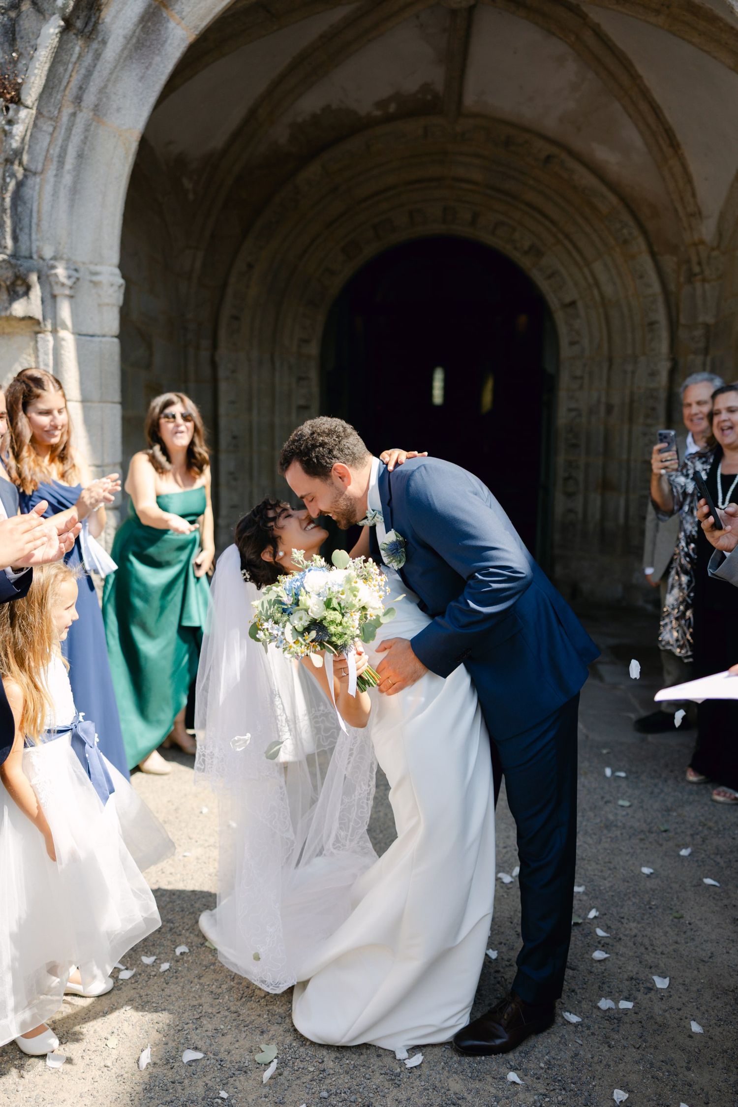 Newlyweds share a romantic kiss outside a stone church arch, surrounded by cheering guests throwing petals.