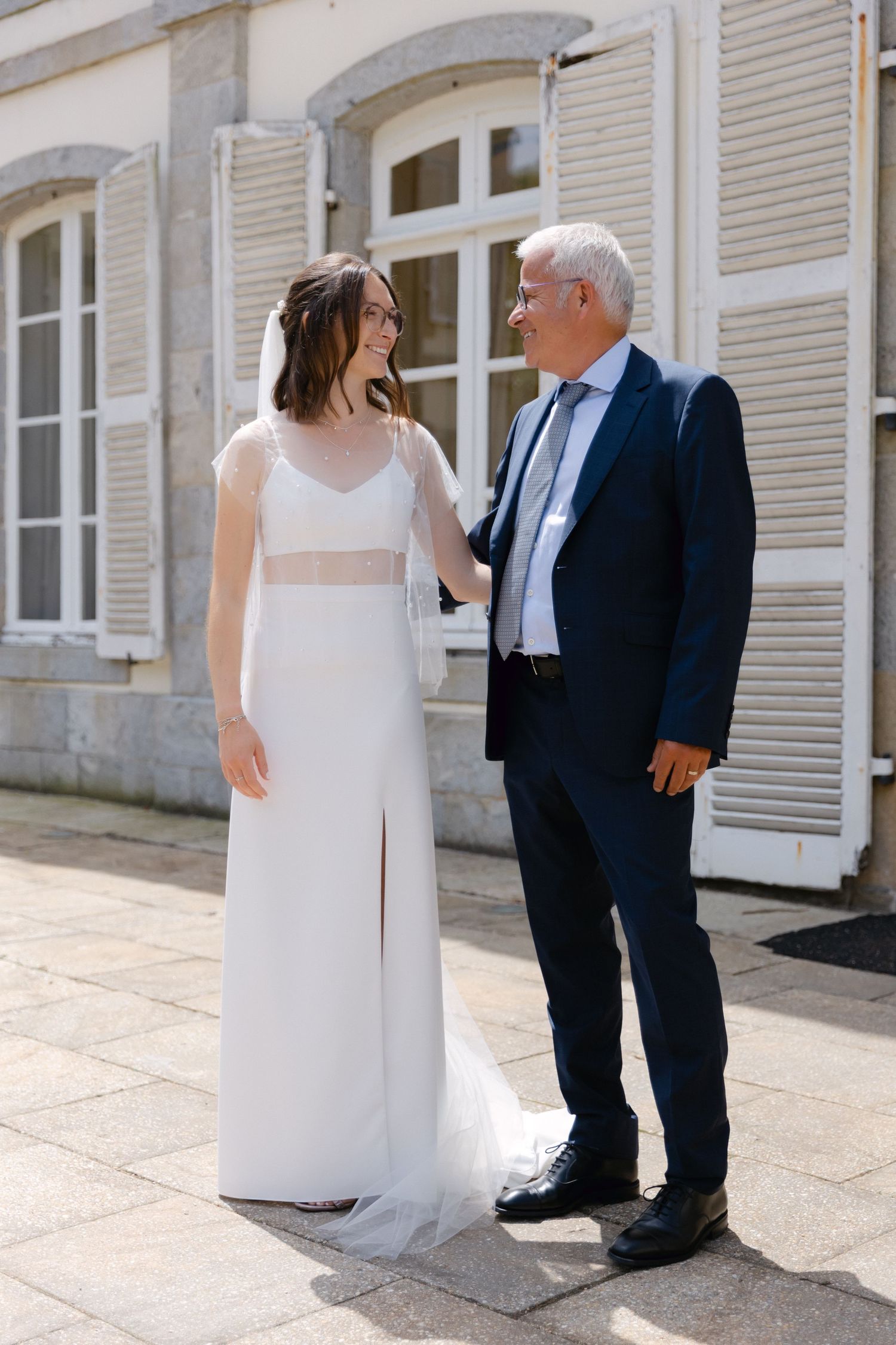 La mariée en robe blanche et l'homme plus âgé en costume bleu marine partagent un sourire à l'extérieur d'un bâtiment français lors d'une jo