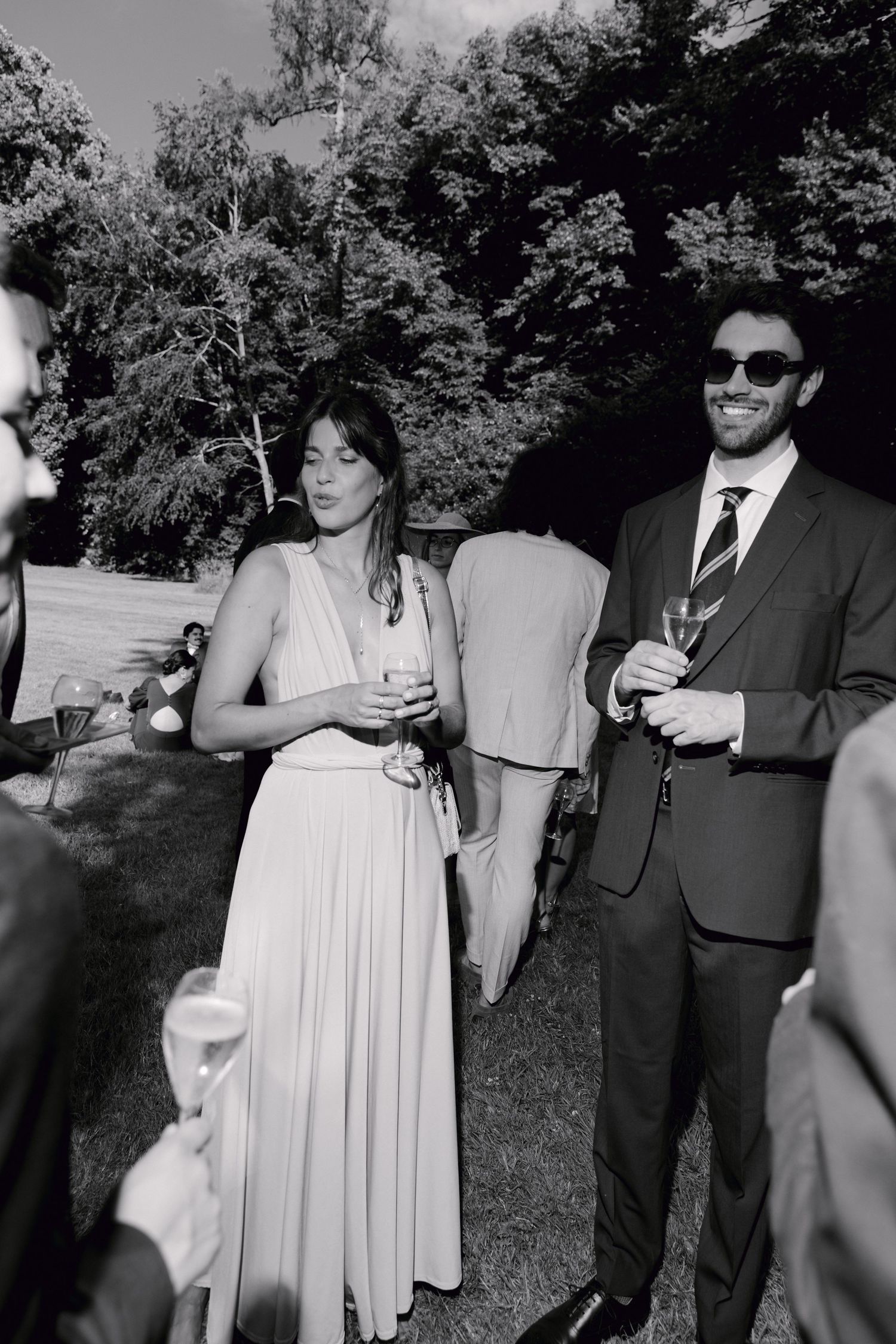 Photo en noir et blanc d'invités de mariage élégamment habillés tenant des verres de champagne à l'extérieur lors d'une réception de jardin.