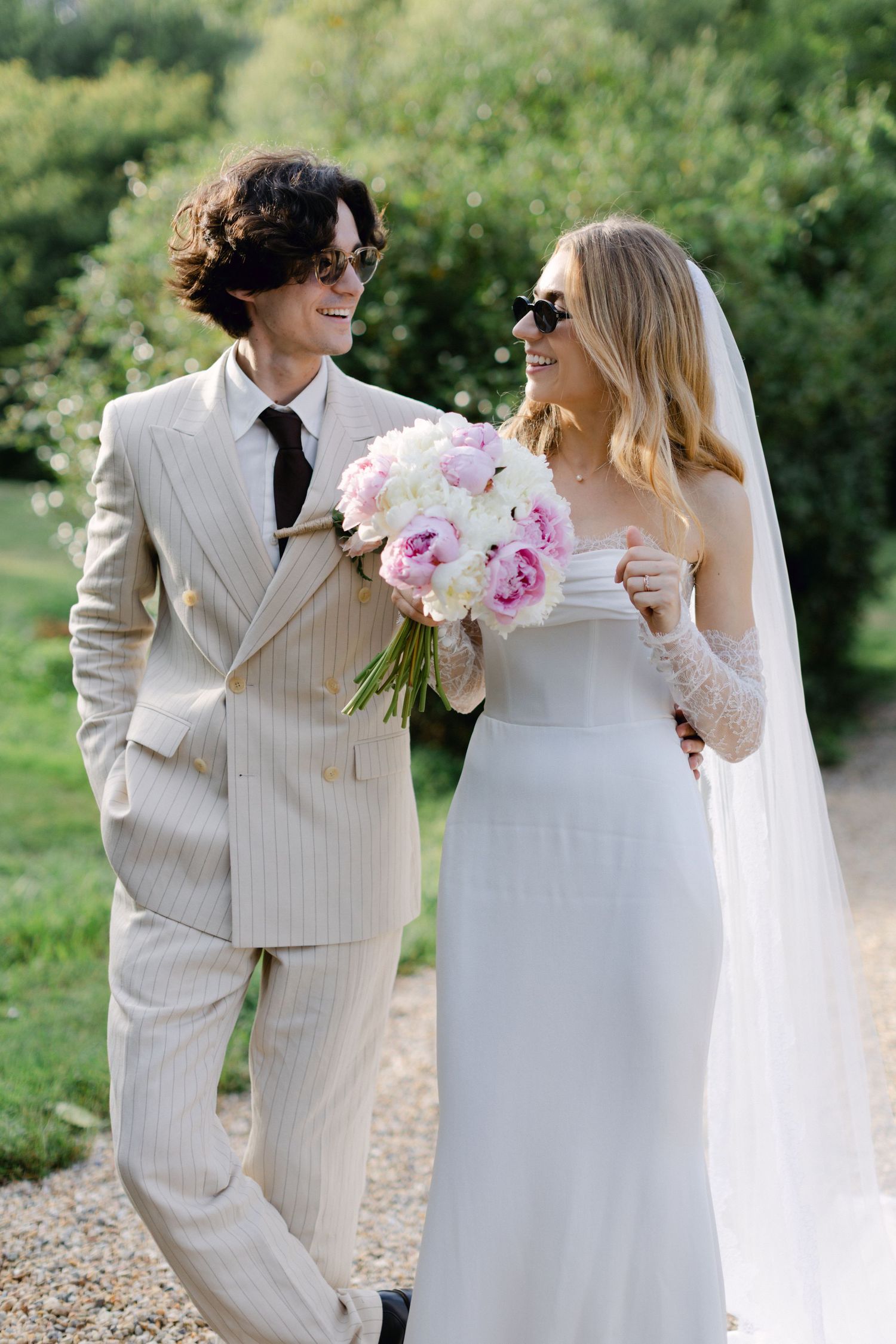 La mariée et le marié souriants en lunettes de soleil se promènent à l'extérieur, elle en voile et robe blanches, lui en costume à rayures.