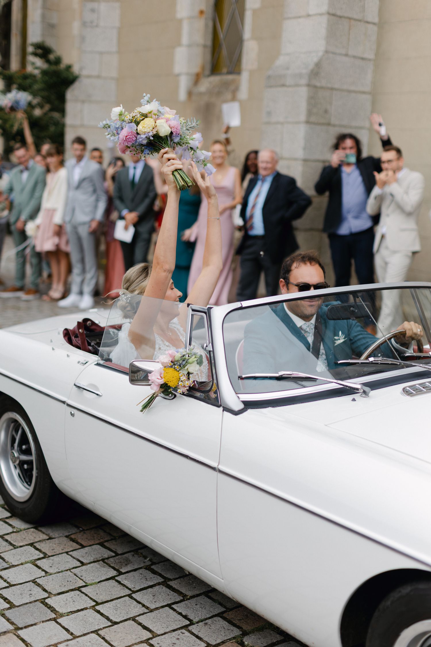 La mariée lève joyeusement un bouquet floral dans une voiture décapotable vintage blanche entourée d'invités au mariage.